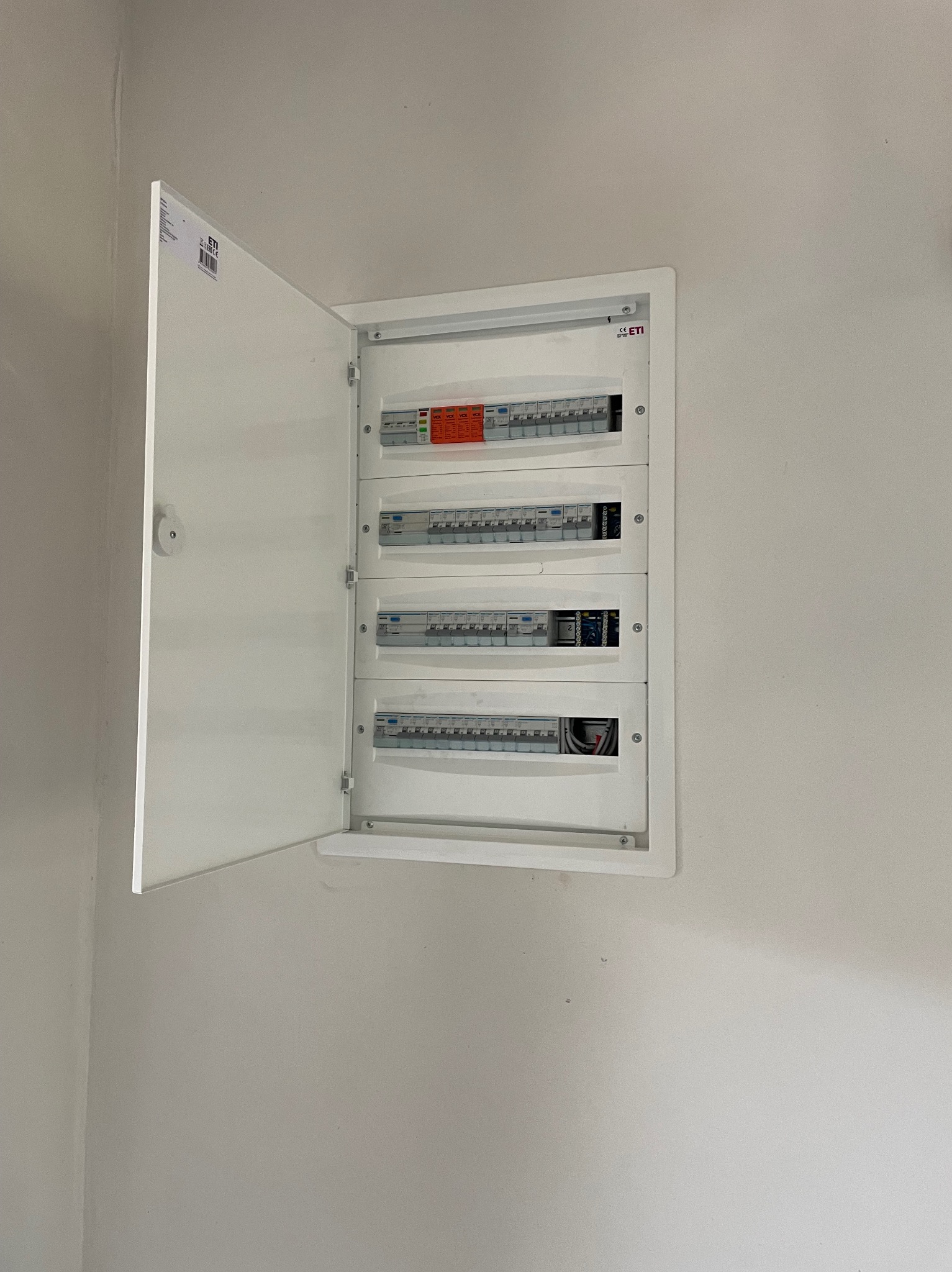 Otwarta, biała skrzynka rozdzielcza z bezpiecznikami na ścianie. Widoczne cztery rzędy bezpieczników. Skrzynka marki ETI.