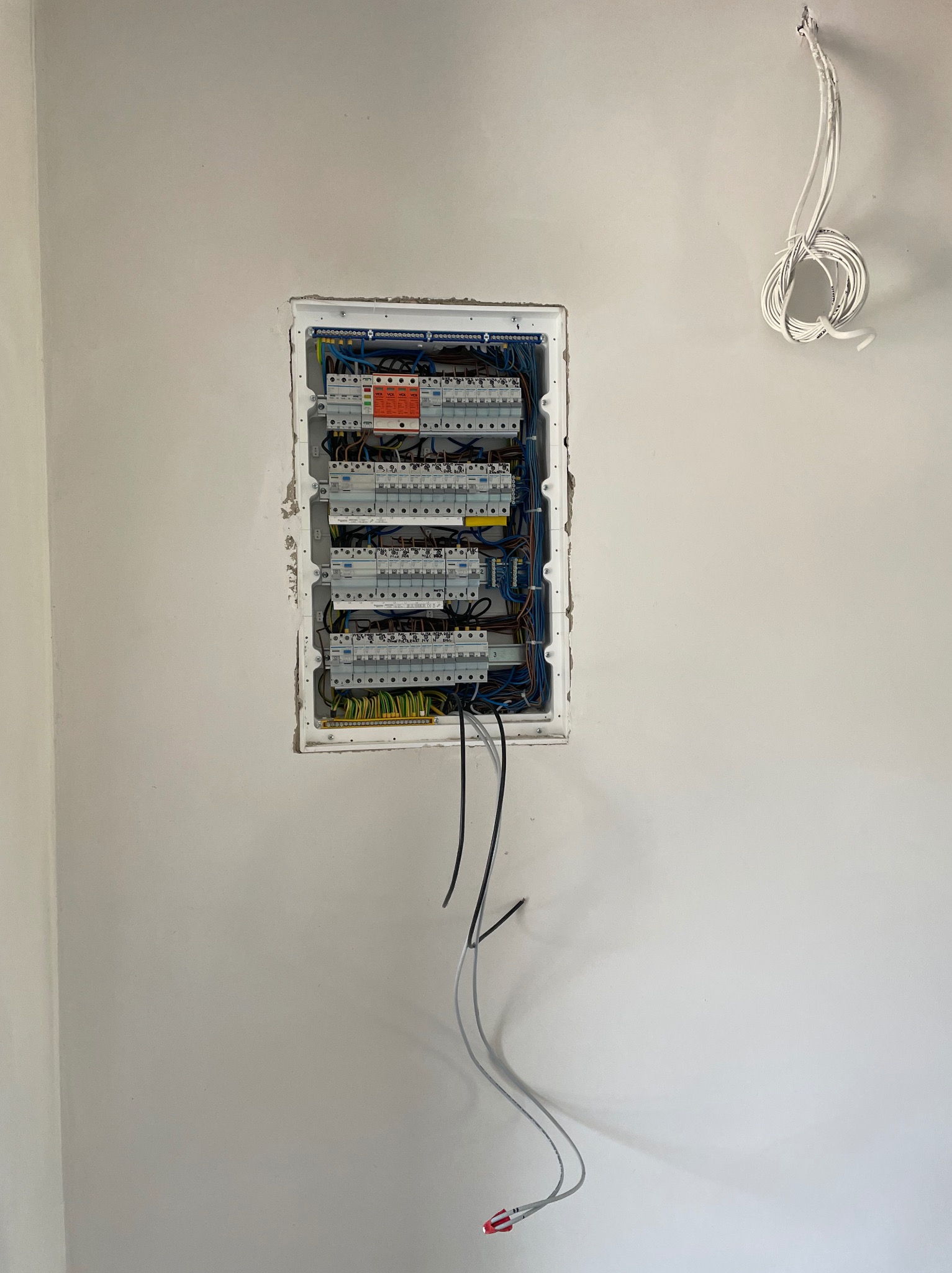 Rozdzielnia elektryczna wnękowa, z widocznymi bezpiecznikami i przewodami, zamontowana na białej ścianie. Dodatkowe przewody zwisają obok.