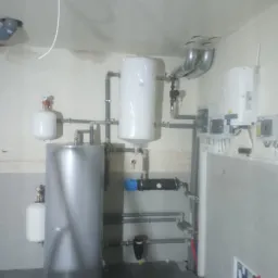 Instalacja centralnego ogrzewania z szarym, cylindrycznym zasobnikiem ciepłej wody, białym zbiornikiem wyrównawczym z manometrem, pompą obiegową z niebieską obudową silnika oraz orurowaniem...