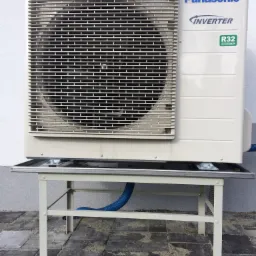 Zewnętrzna jednostka klimatyzacji Panasonic Inverter R32 zamontowana na metalowym stelażu na tle szarej ściany i brukowanej powierzchni.