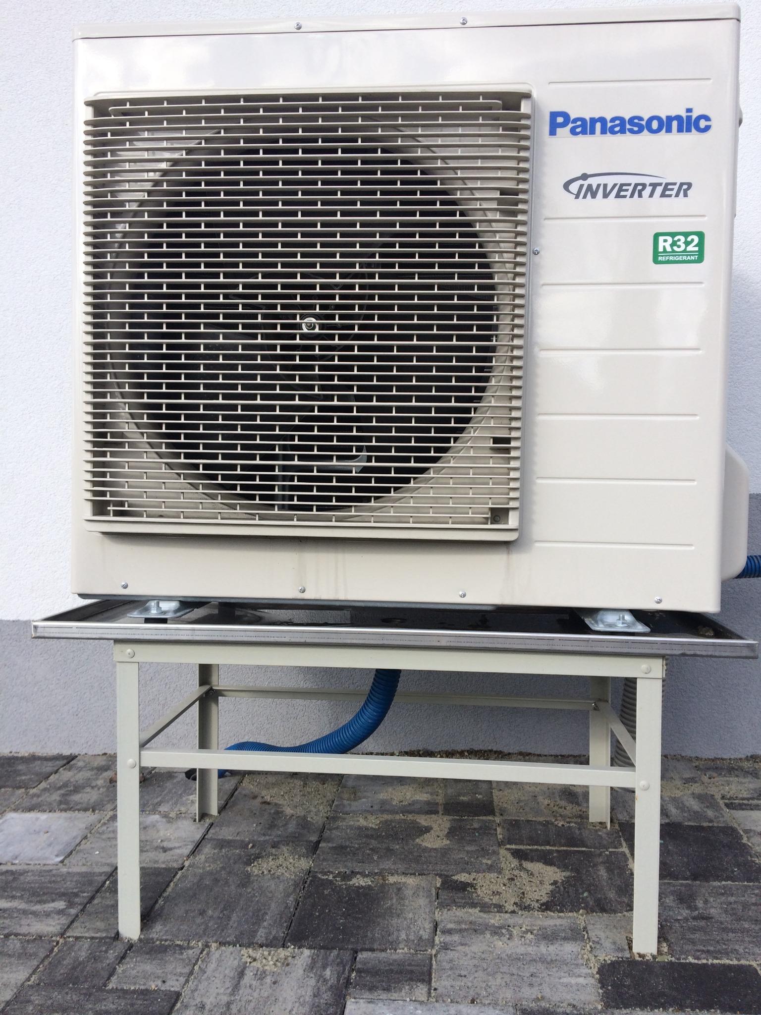 Zewnętrzna jednostka klimatyzacji Panasonic Inverter R32 zamontowana na metalowym stelażu na tle szarej ściany i brukowanej powierzchni.