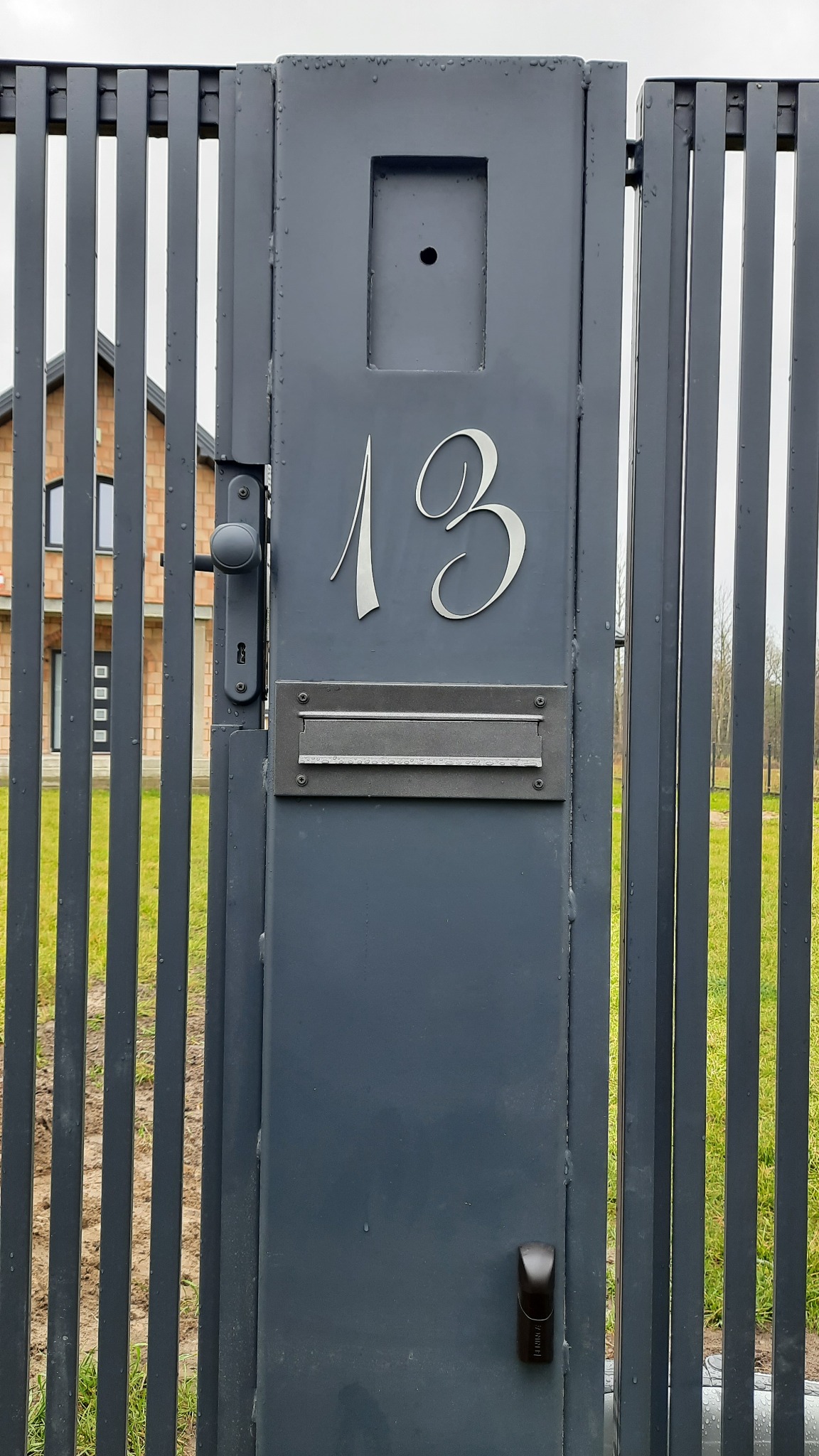 Nowoczesna, szara brama metalowa z numerem domu '13' wykonanym ozdobną czcionką i skrzynką na listy, widoczny fragment domu w tle.