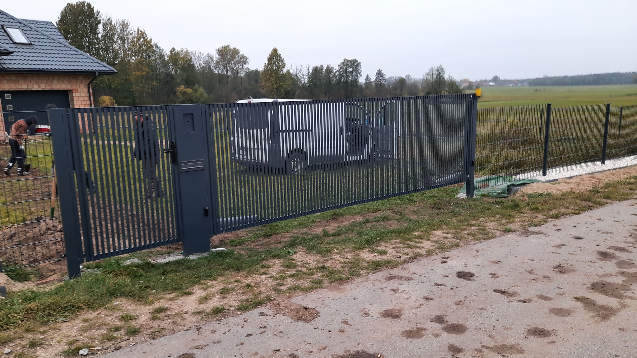 Nowoczesne, grafitowe ogrodzenie panelowe z furtką i skrzynką na listy przed domem w trakcie montażu, widoczni pracownicy i częściowo zasłonięty samochód dostawczy.