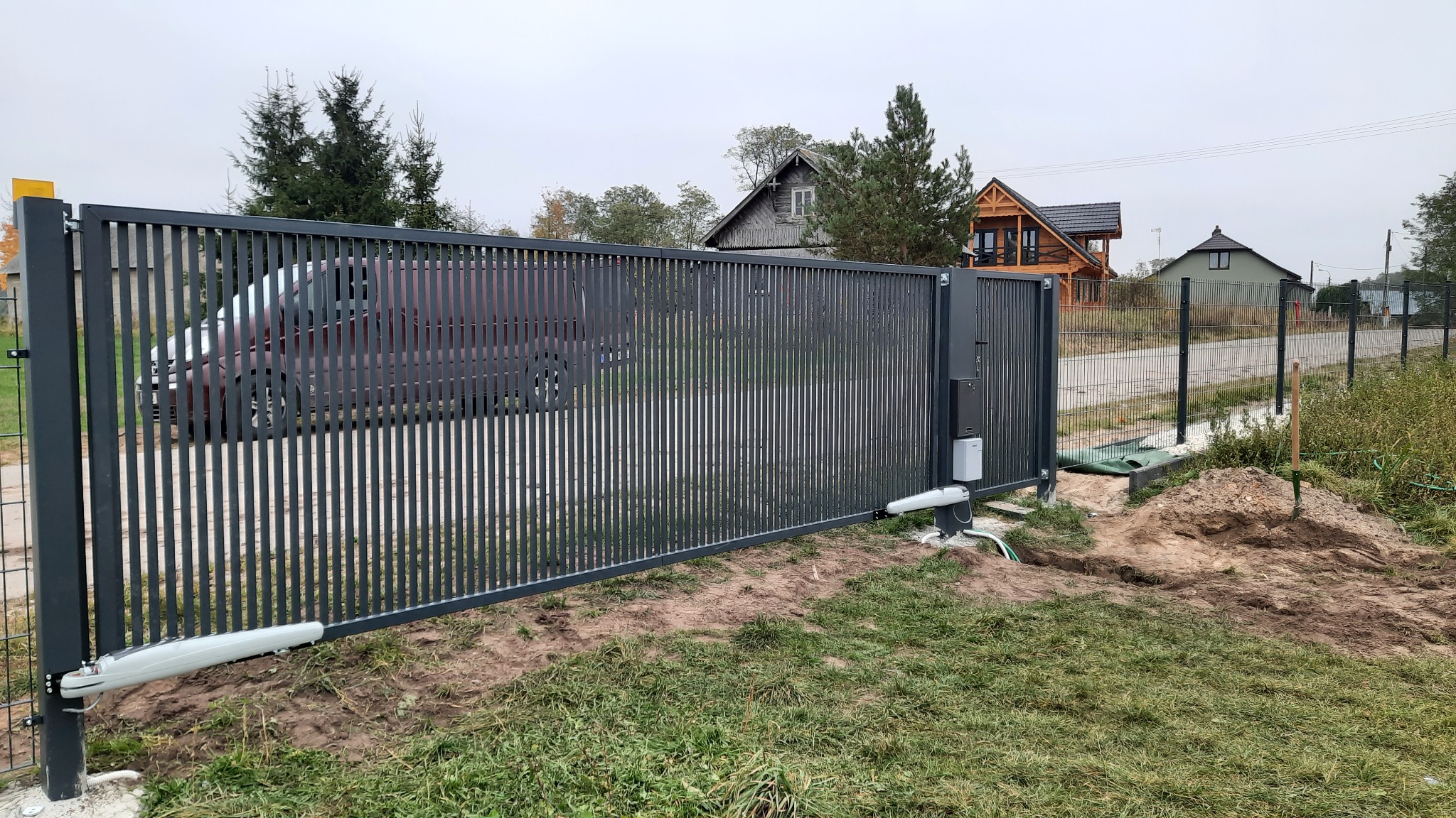 Nowoczesna, przesuwna brama metalowa z automatyką, widoczna na tle domów jednorodzinnych i częściowo zakrywająca zaparkowany samochód, z widocznym wykopem po instalacji.
