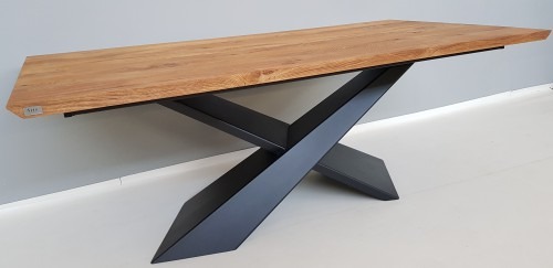 Stół z blatem z jasnego drewna i czarną, metalową podstawą w kształcie litery X, ustawiony na białej podłodze, z szarą ścianą w tle.