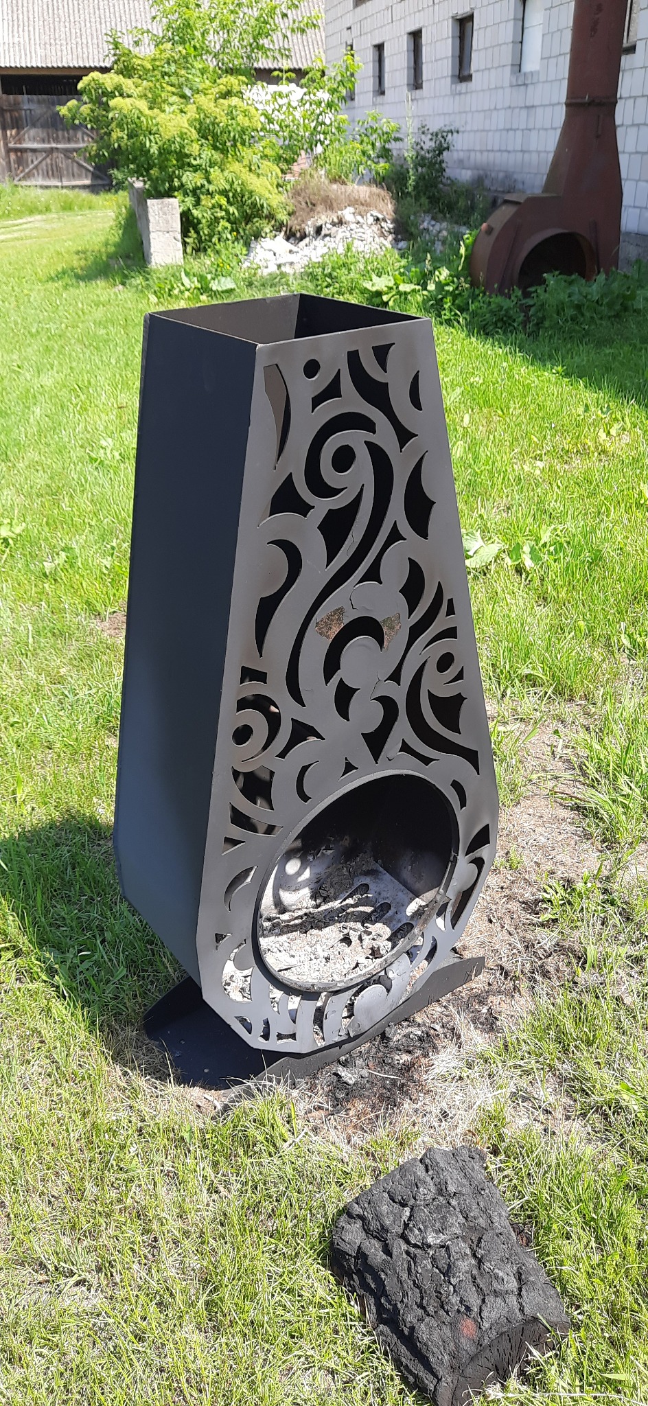 Metalowy kosz na ogień z dekoracyjnymi wycięciami, stojący na trawie, z widocznymi resztkami popiołu w środku i kawałkiem spalonego drewna obok.