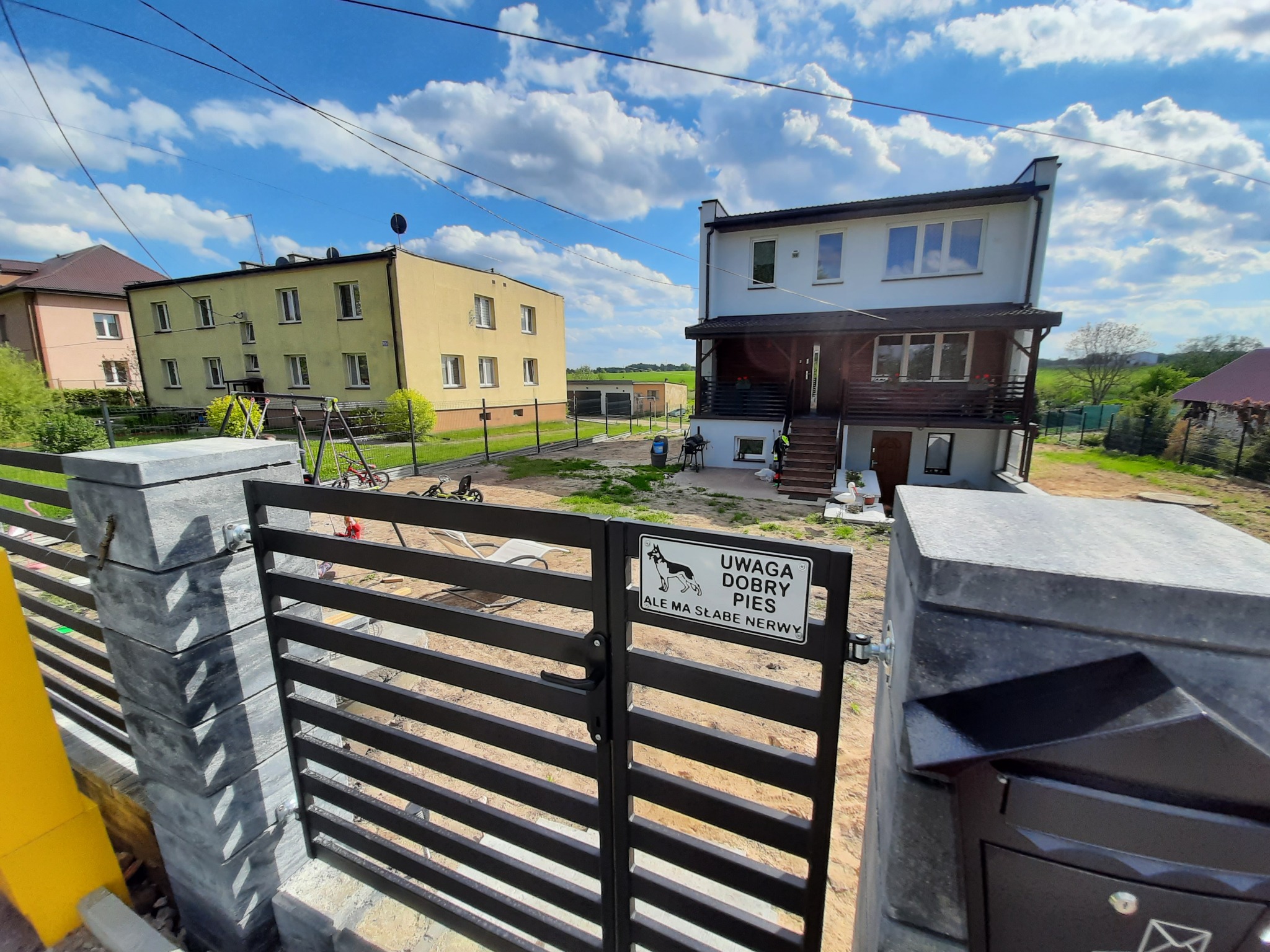 Nowoczesna, pozioma, metalowa brama wjazdowa z szarymi, betonowymi słupkami i tabliczką ostrzegawczą 'Uwaga dobry pies, ale ma słabe nerwy' na tle dwupiętrowego domu i błękitnego nieba z chmurami.