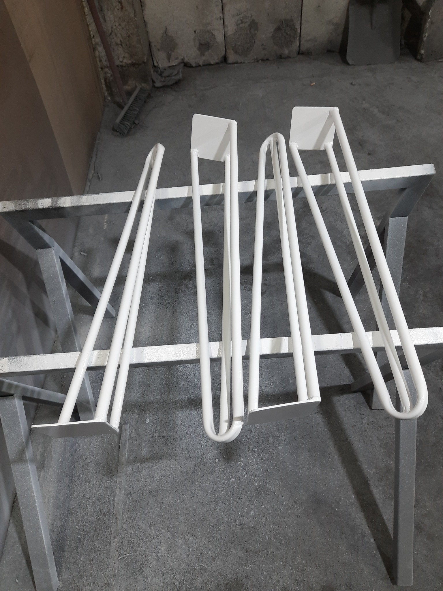 Białe, świeżo pomalowane proszkowo metalowe nogi do stołu o różnym kształcie, suszące się na metalowym stojaku w warsztacie.