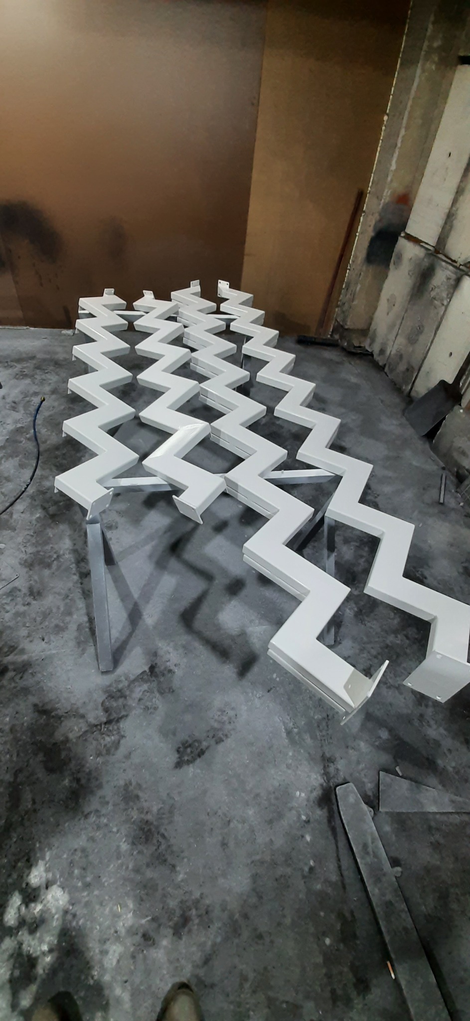 Białe, zygzakowate elementy metalowe, prawdopodobnie fragmenty balustrady lub ogrodzenia, ułożone na szarej betonowej podłodze w warsztacie, widoczne narzędzia i ślady obróbki.