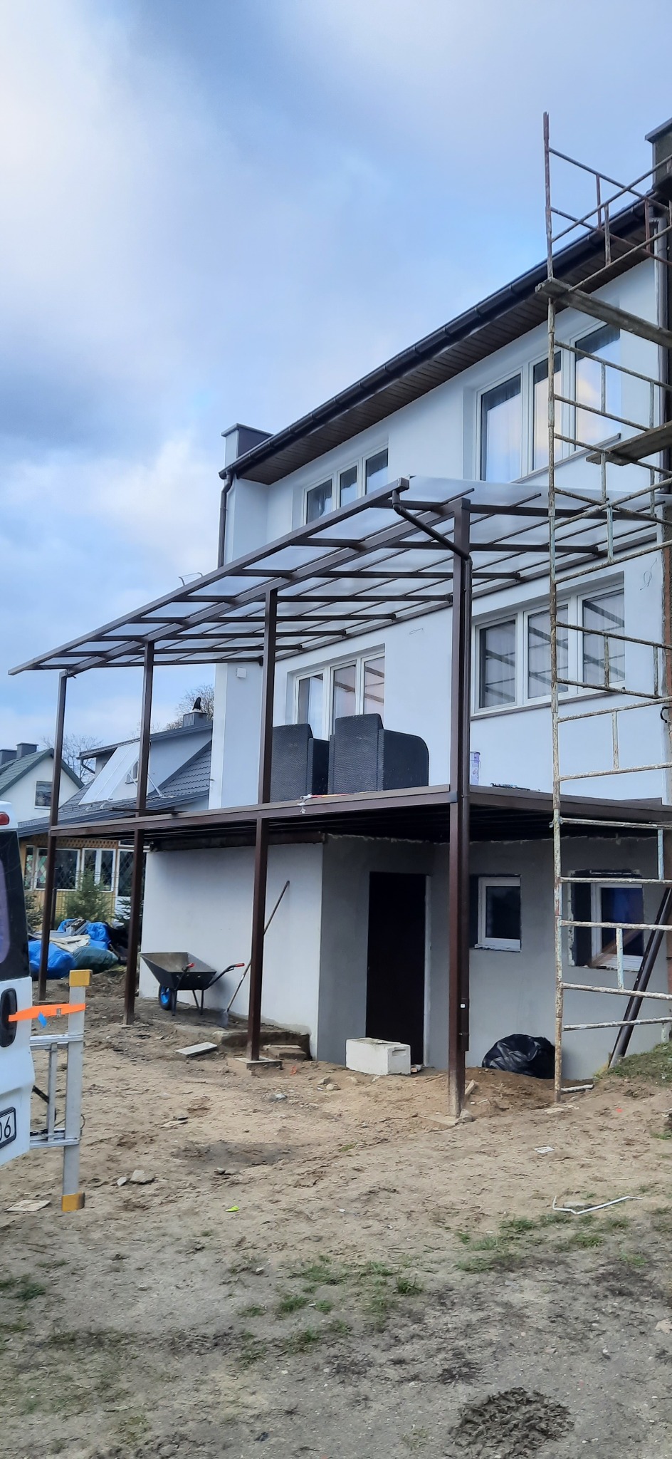 Zadaszenie tarasu z metalową konstrukcją przy dwupiętrowym domu, widoczne rusztowanie i elementy budowlane na ziemi.