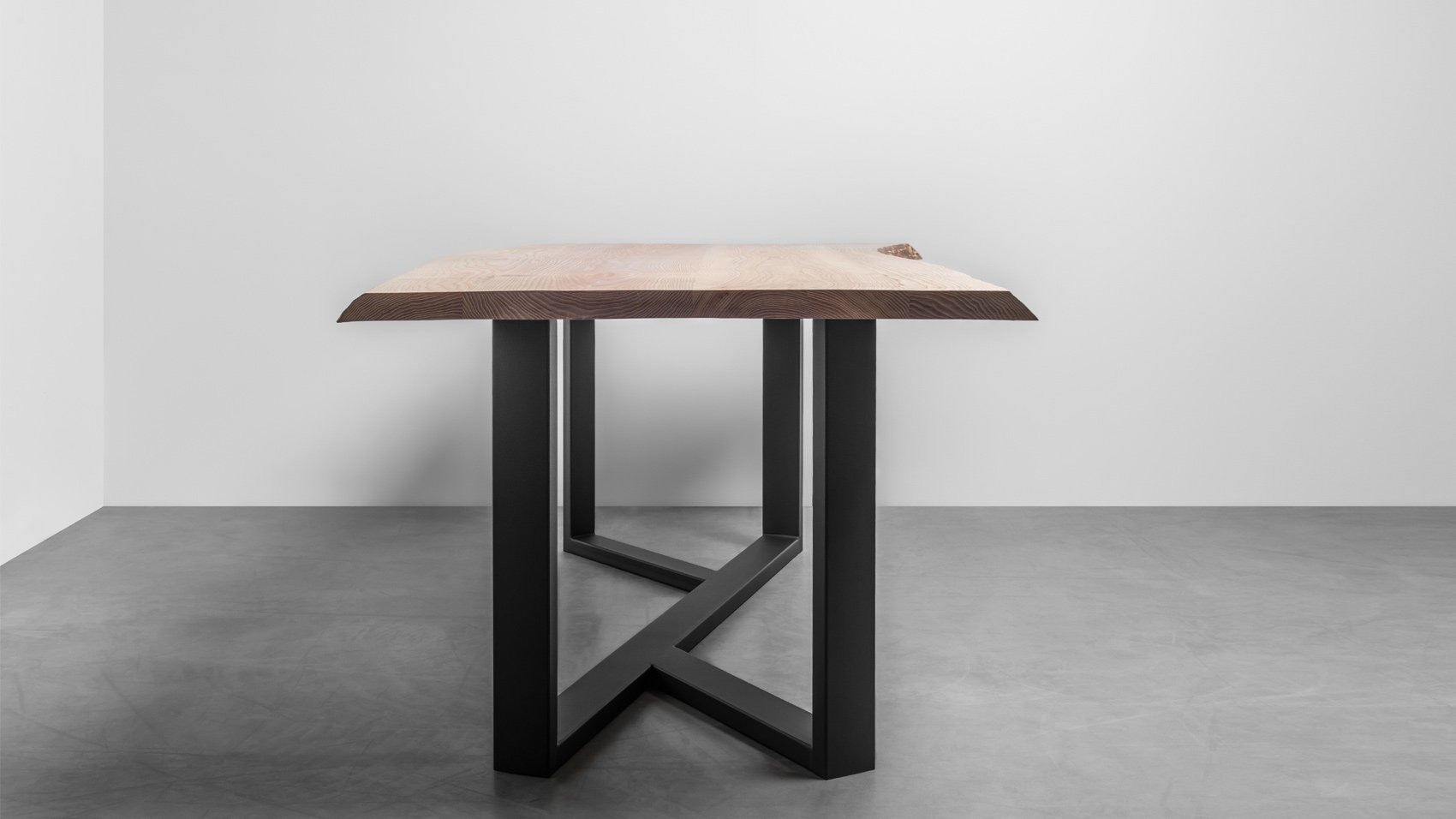 Stół z blatem z jasnego drewna o naturalnej krawędzi i czarnymi, metalowymi nogami o geometrycznym kształcie na szarym tle.