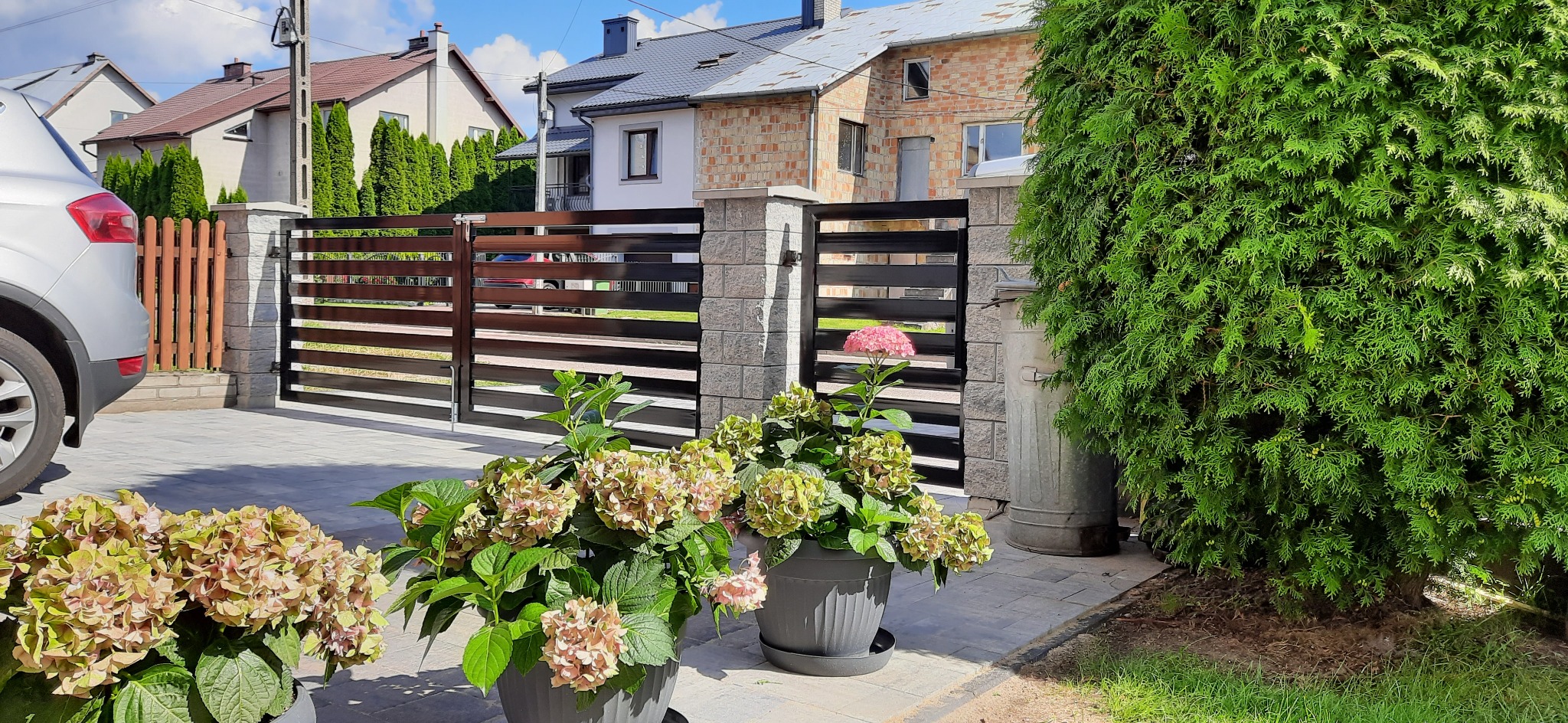 Nowoczesna brama posesyjna z poziomymi przęsłami w kolorze ciemnego brązu, osadzona na słupkach z szarej kostki, z widocznymi donicami z hortensjami i fragmentem samochodu w tle.