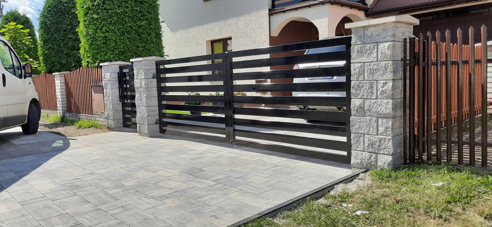 Nowoczesna brama wjazdowa z czarnymi, poziomymi przęsłami, osadzona na murowanych z jasnych bloczków słupkach, widok z zewnątrz na podjazd wyłożony szarą kostką brukową.