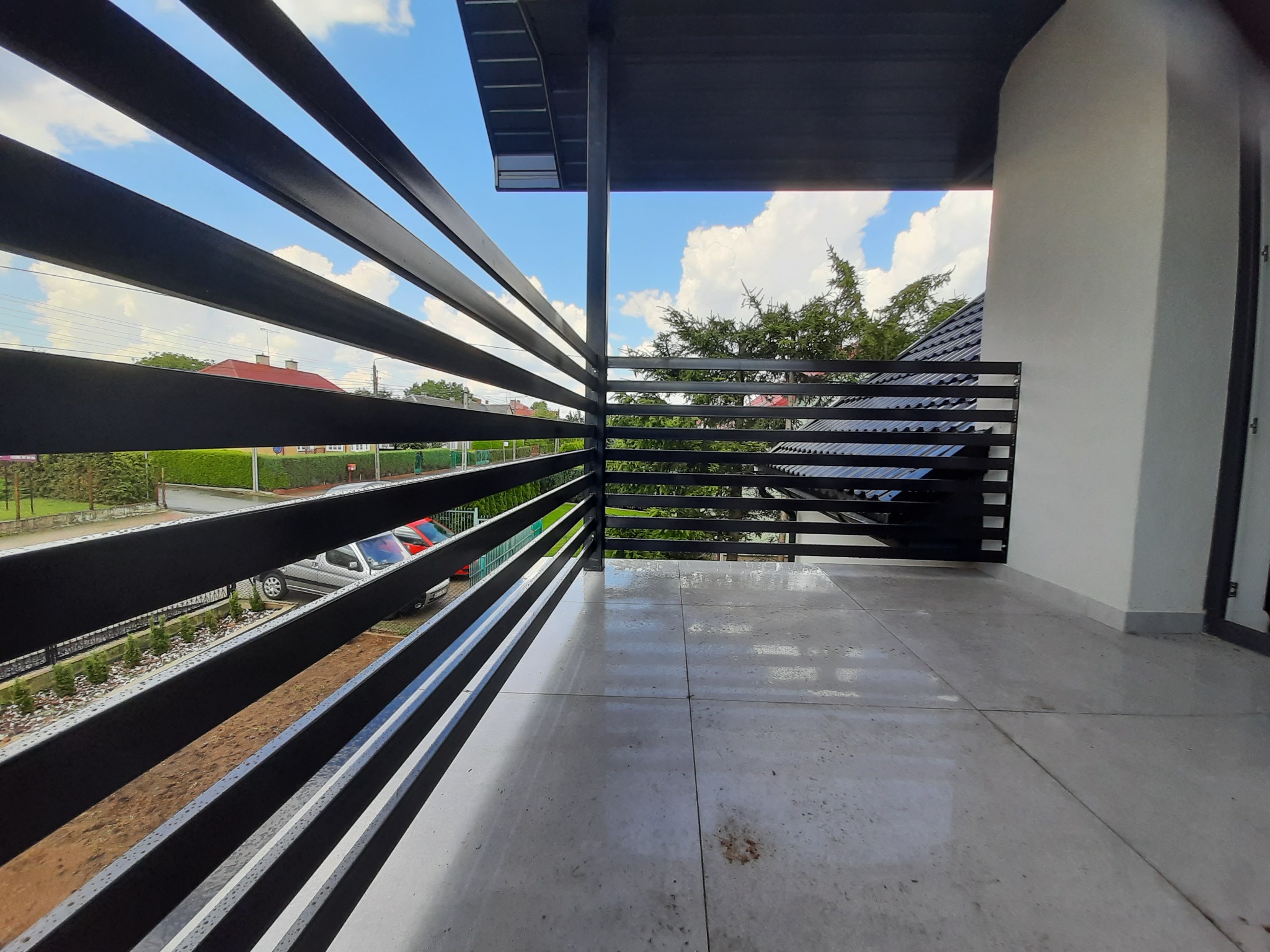 Nowoczesna, czarna balustrada balkonowa z metalowymi poziomymi prętami, widok na okolicę z fragmentem nieba i drzew, szara płytka podłogowa.