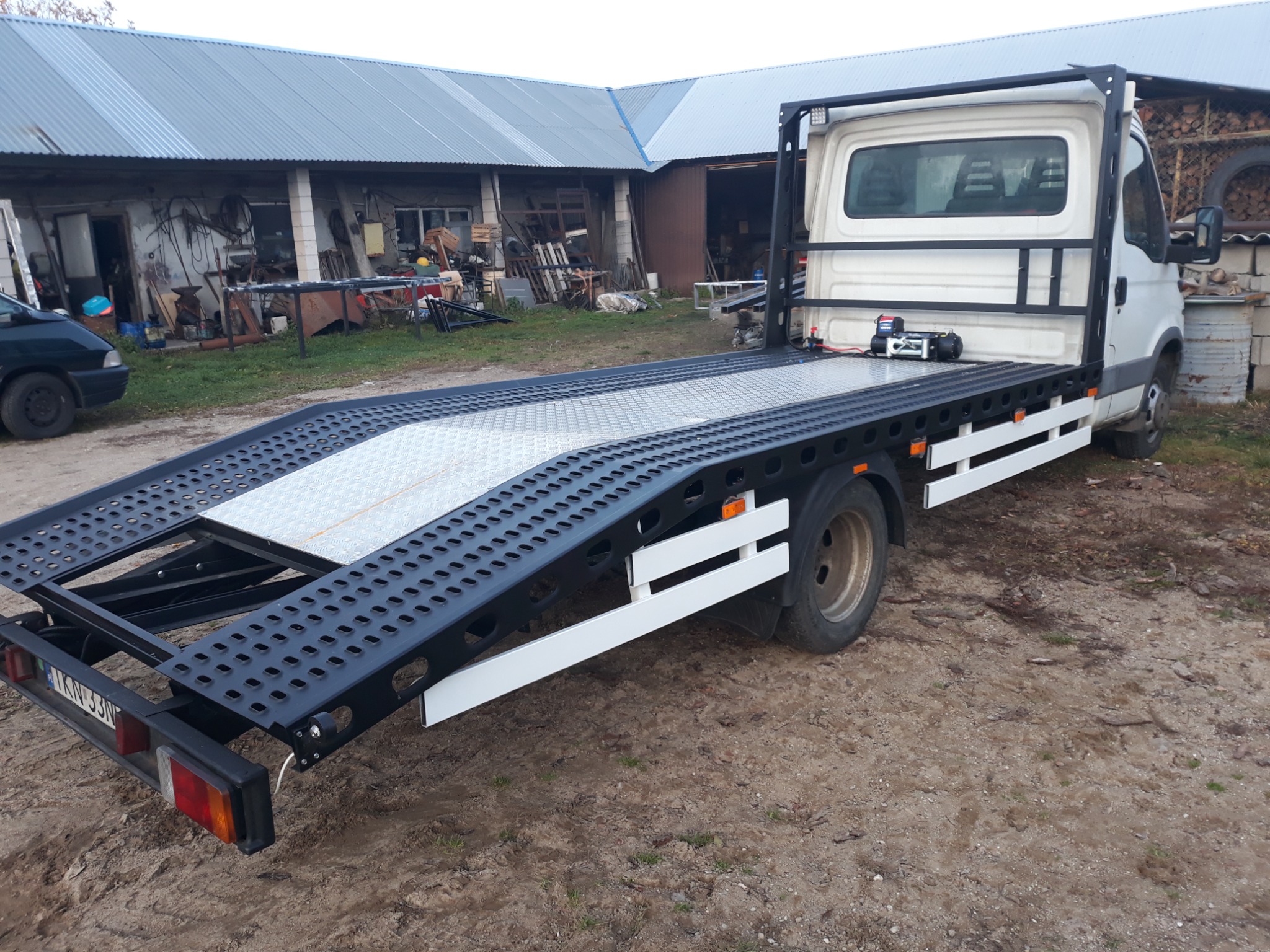 Laweta samochodowa z najazdami, pomalowana na czarno-biało, zaparkowana na ziemi przed budynkiem gospodarczym z metalowym dachem.