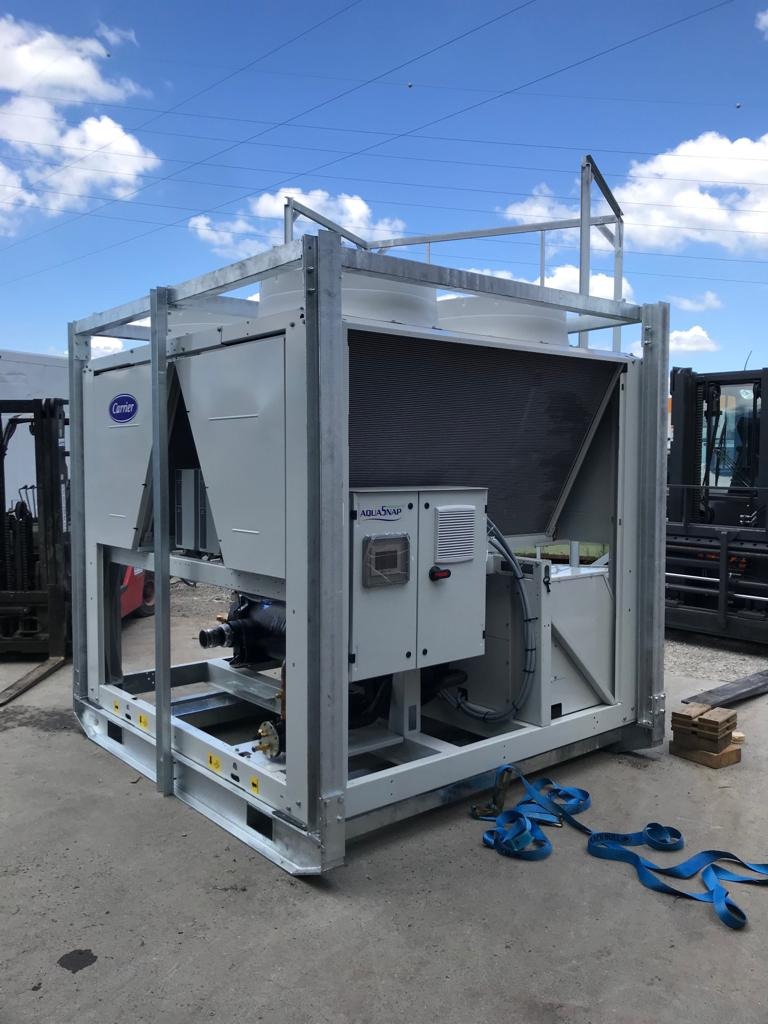 Kompaktowa przemysłowa chłodnica Carrier AquaSnap w metalowej ramie, widoczne elementy instalacji, na zewnątrz, w tle wózki widłowe i błękitne niebo.