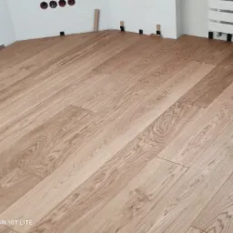 Jasne panele podłogowe ułożone w rogu pomieszczenia, widoczne listwy dystansowe przy ścianach i grzejnik.