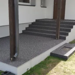 Zewnętrzny taras i schody wejściowe pokryte szarą, drobnoziarnistą posadzką żywiczną, z widoczną rynną odprowadzającą wodę deszczową, przy białej ścianie domu.