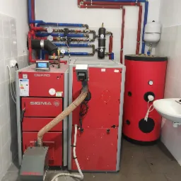 Kompletna instalacja kotłowni z dwoma kotłami Defro, zasobnikiem ciepłej wody i osprzętem, widoczne rury w kolorach czerwonym i niebieskim oraz biała umywalka.