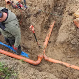Wykop z ułożonymi pomarańczowymi rurami kanalizacyjnymi, widoczny fragment niebieskiej rury, łopata wbita w ziemię, narzędzia elektryczne leżące na ziemi w pobliżu wykopu, pracownik w roboczym...