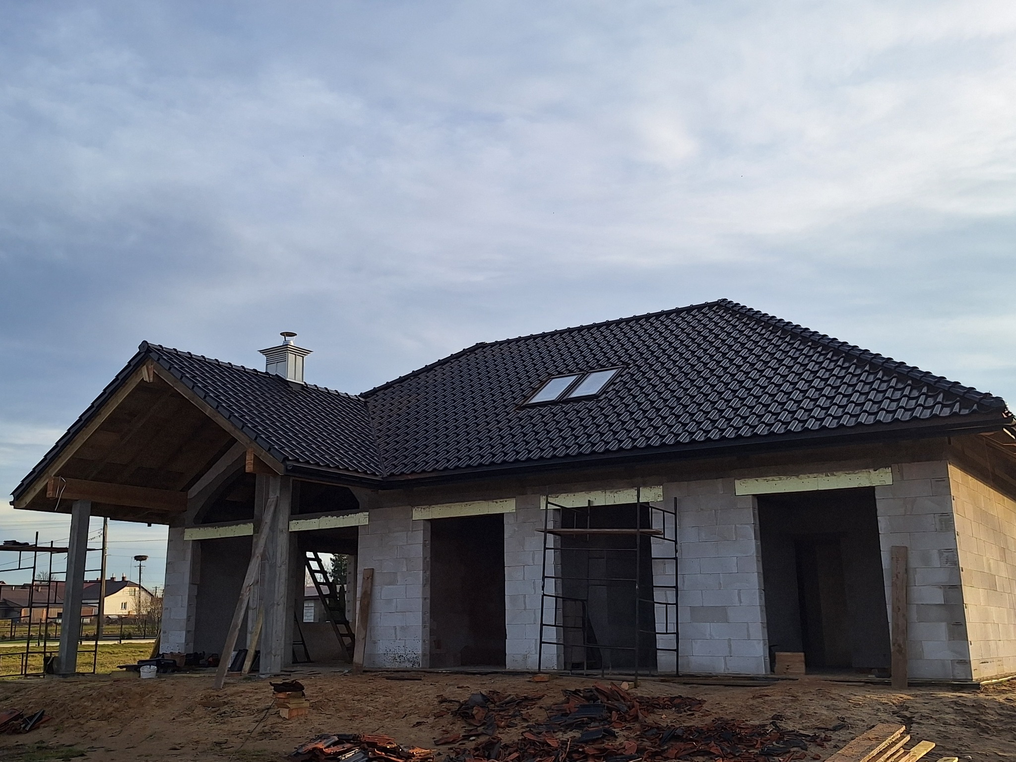 Dom w trakcie budowy z ciemnym dachem i oknem dachowym. Widoczne bloczki betonowe, rusztowania i elementy drewniane konstrukcji. Częściowo ułożona dachówka.