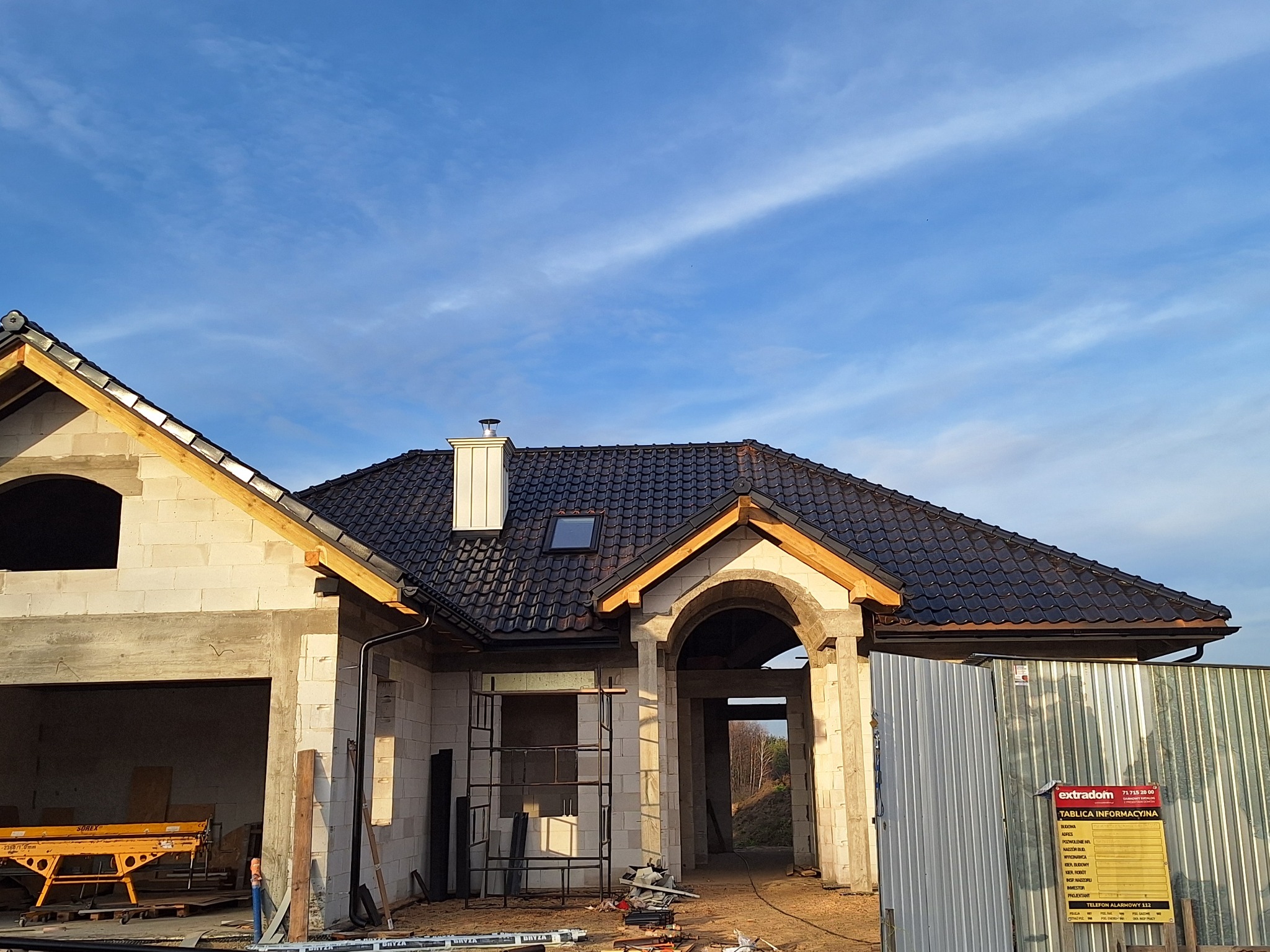Dom w trakcie budowy z ciemną dachówką, widoczne rusztowania i tablica informacyjna. Częściowo wykończony dach z oknem dachowym i kominem. Błękitne niebo w tle.