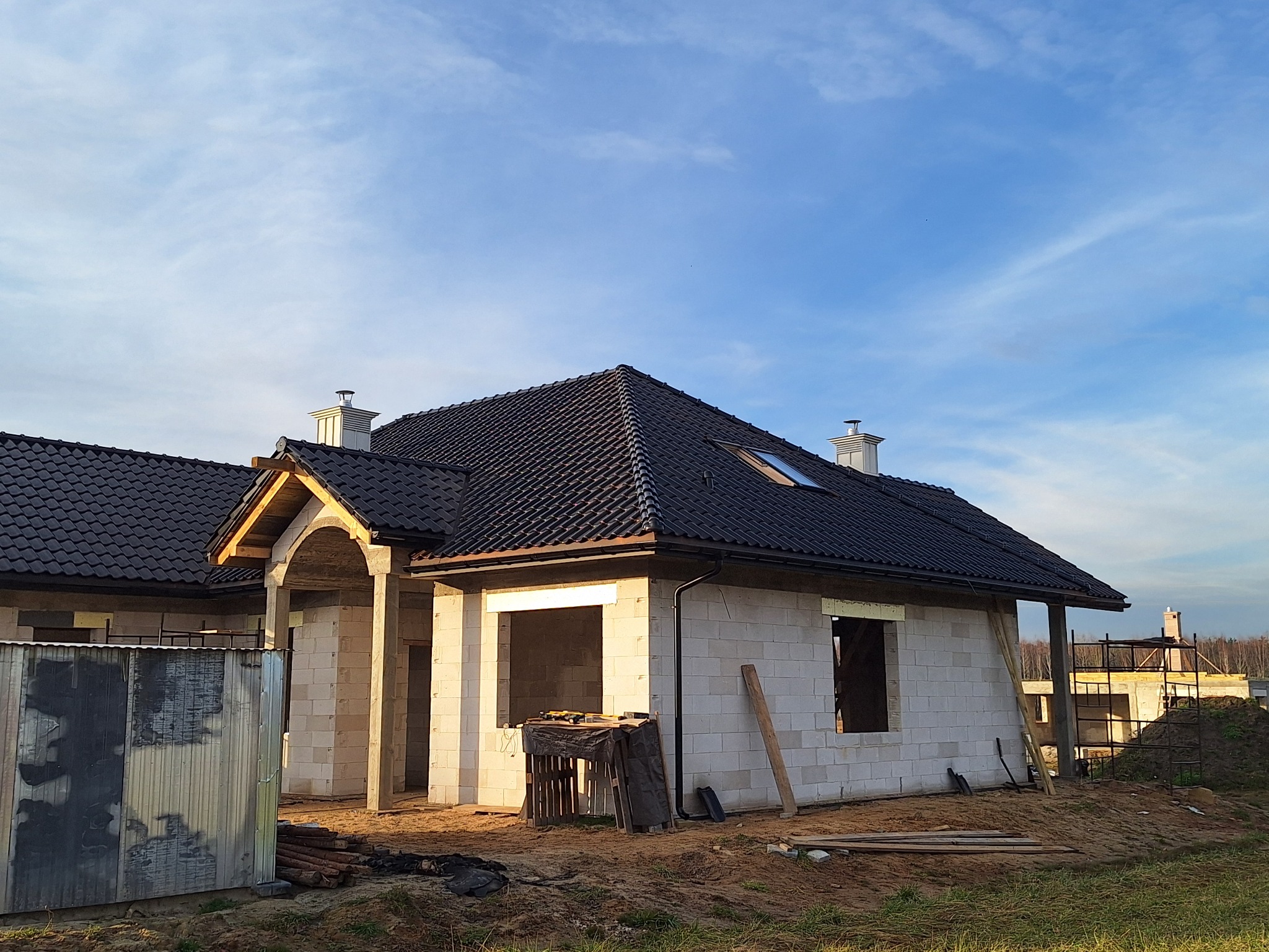 Dom w trakcie budowy z czarnym dachem i widocznymi oknami. Budynek z białych pustaków, otoczony ziemią i trawą. Częściowo ukończony dach z lukarną.