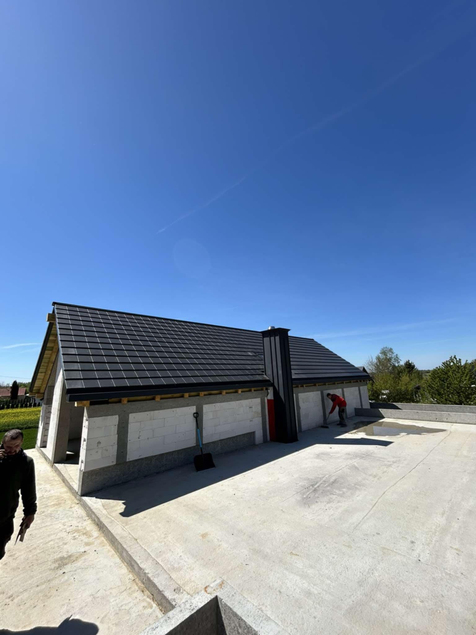 Dom w trakcie budowy z czarnym dachem z dachówki, widoczny komin i pracownik w czerwonej koszulce. Ściany z białego betonu komórkowego.