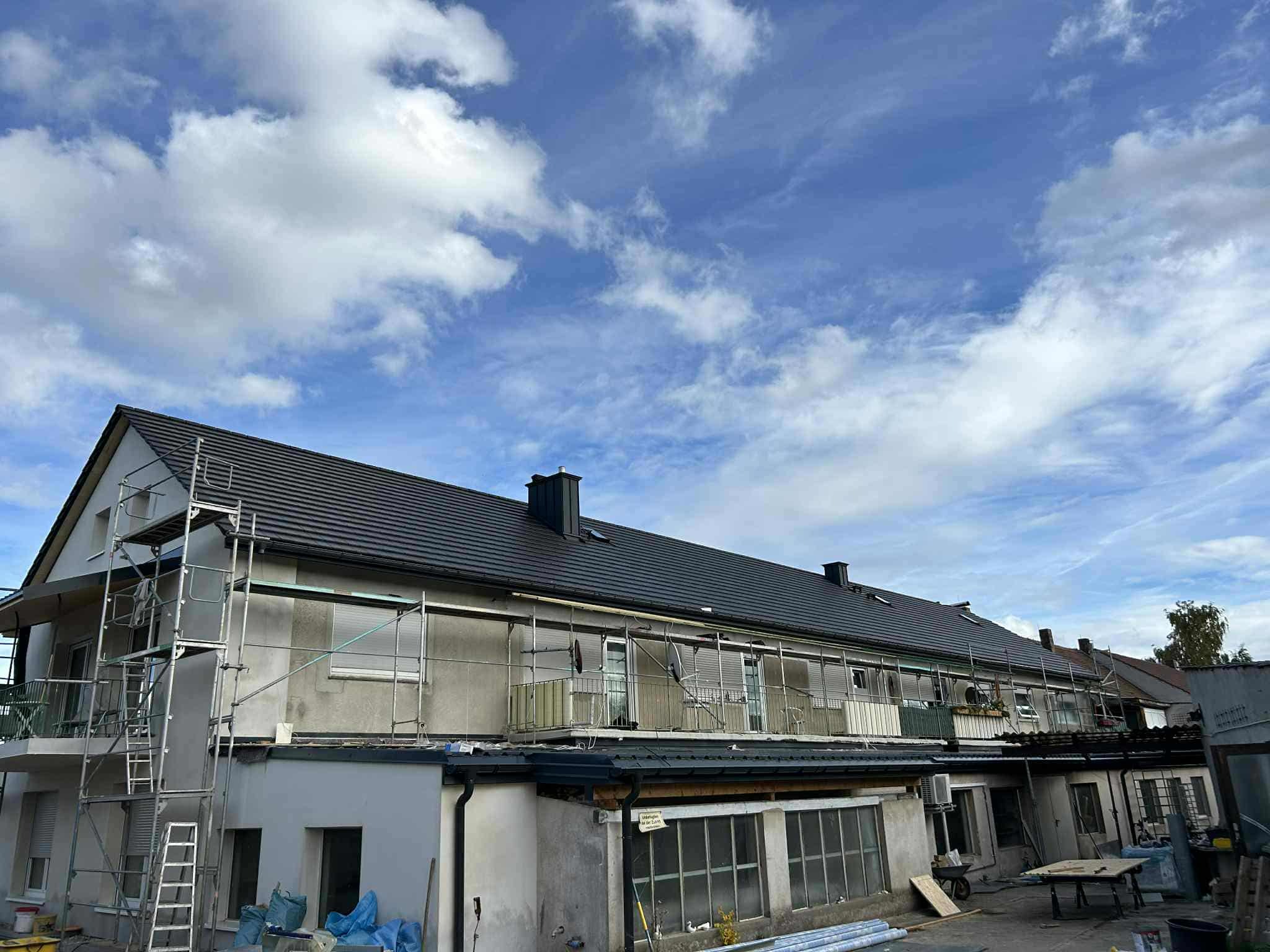 Budynek w trakcie prac remontowych z rusztowaniem, widoczne nowe, ciemne pokrycie dachowe i fragment nieba z chmurami. Częściowo otynkowana elewacja.