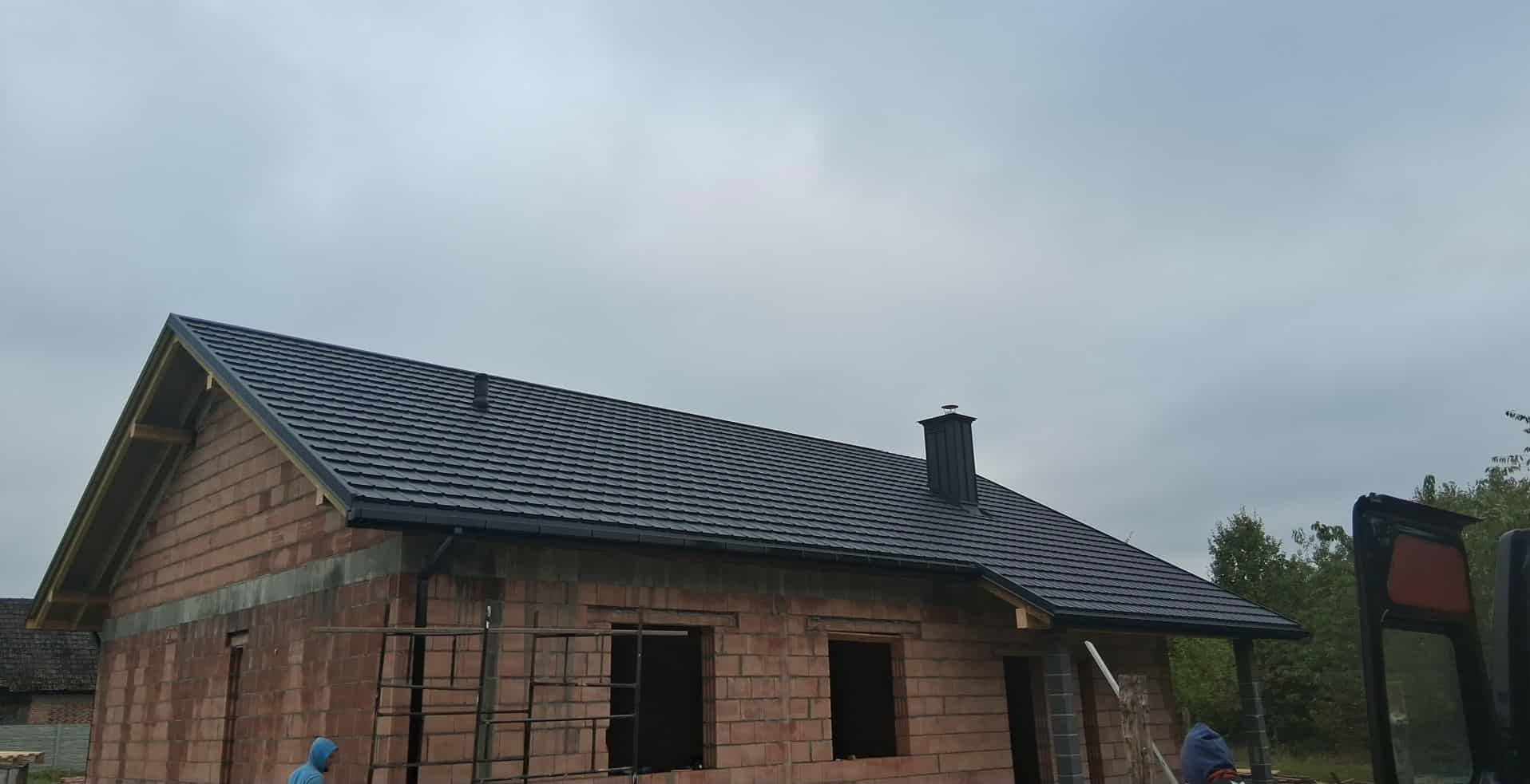 Dom w trakcie budowy z nowym, ciemnym dachem z dachówki ceramicznej i kominem. Widoczne rusztowania i niezabudowane okna. Pochmurne niebo w tle.
