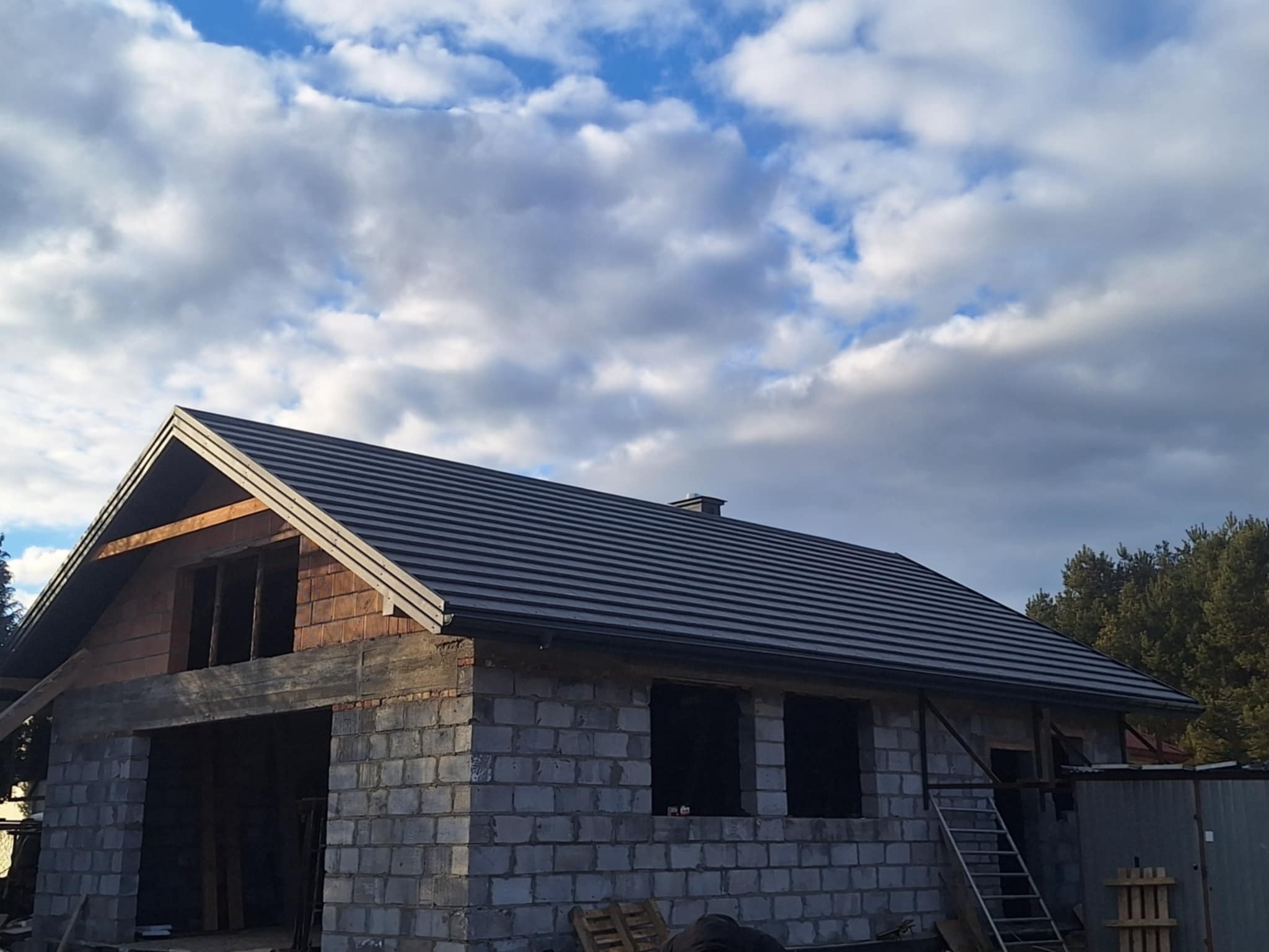 Szary dach z blachy na surowym budynku w trakcie budowy, widoczne pustaki i drewniana konstrukcja. W tle błękitne niebo z chmurami.