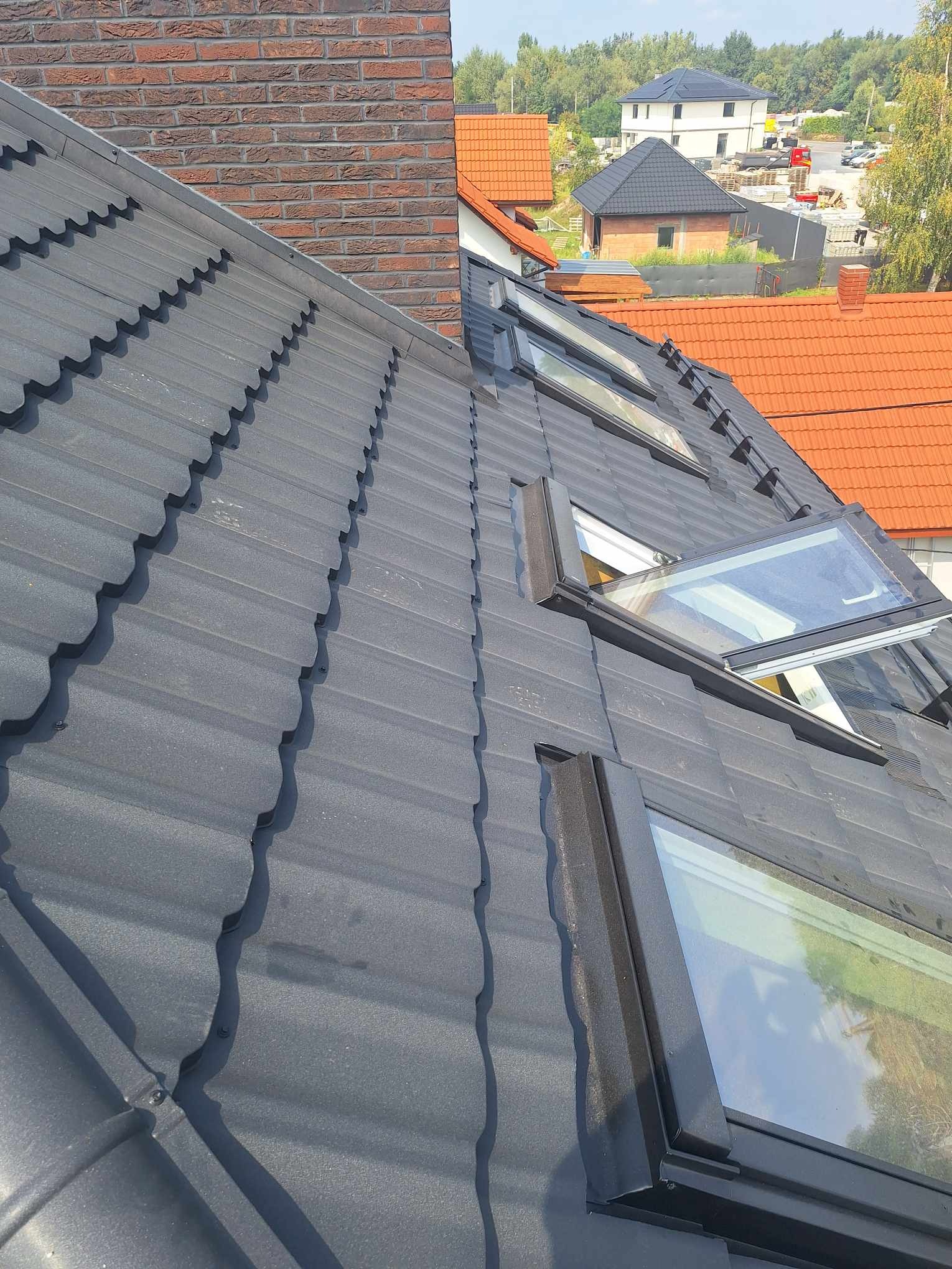 Grafitowy dach z oknami połaciowymi, widok z góry, ceglany komin i zabudowa w tle. Nowoczesne pokrycie dachowe z wyraźną fakturą i detalami wykończenia.