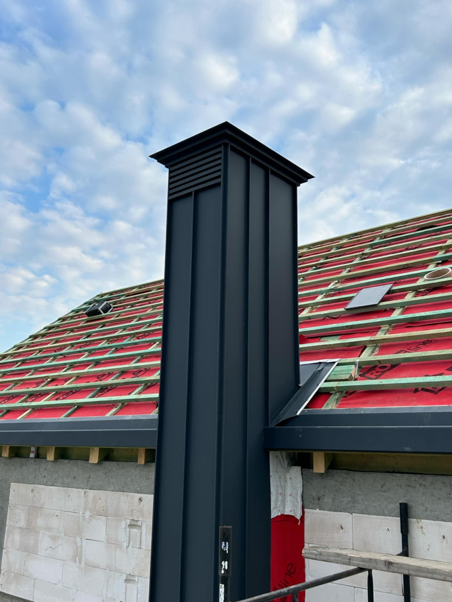 Nowoczesny, grafitowy komin z blachy na tle dachu w trakcie budowy z czerwoną membraną i drewnianą konstrukcją. Widoczne białe bloczki fundamentowe.