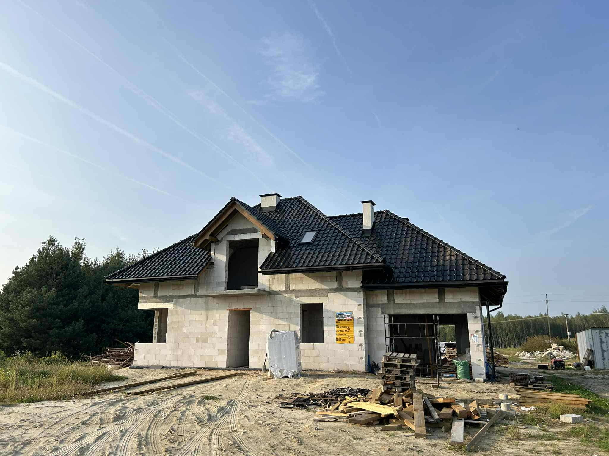 Dom jednorodzinny w trakcie budowy, z czarną dachówką i widoczną konstrukcją z bloczków. Na pierwszym planie materiały budowlane i teren budowy.