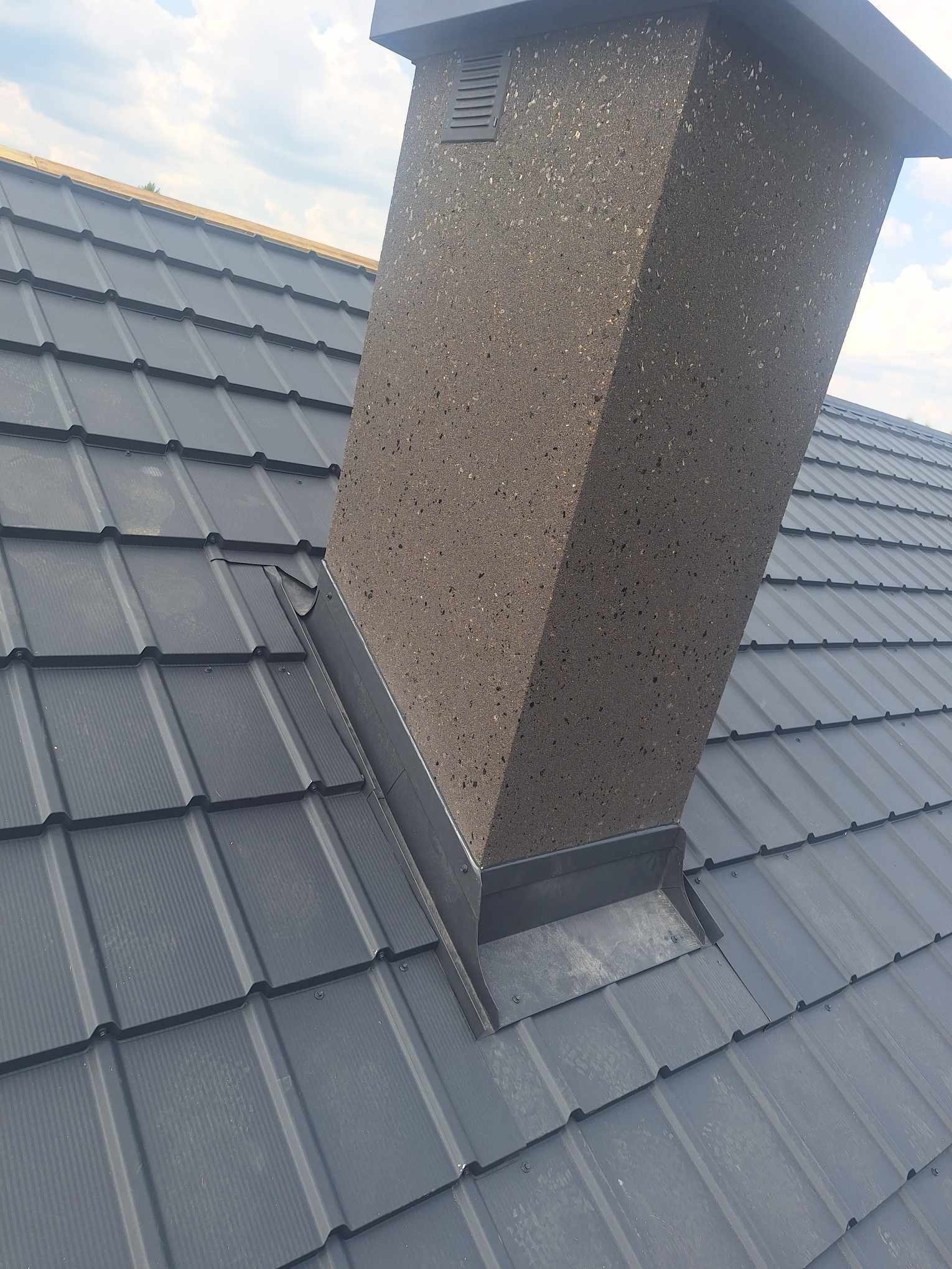 Szary dach z blachy trapezowej z kominem o szorstkiej fakturze, widok z góry, oświetlenie dzienne, detale łączeń blachy i obróbek komina.