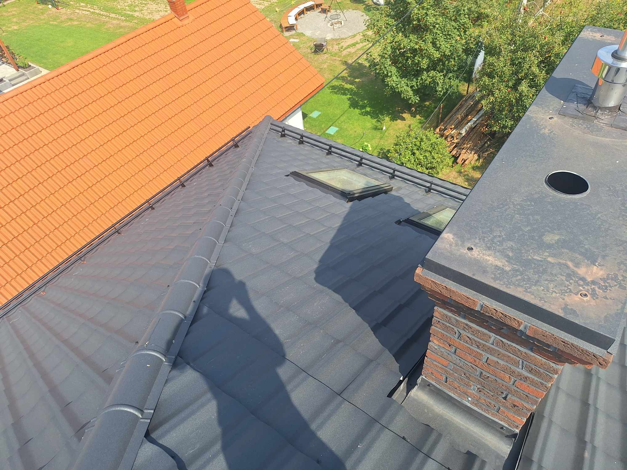 Grafitowy dach z dachówką modułową, widoczne okna dachowe i komin z cegły klinkierowej. W tle fragment dachu z pomarańczowej dachówki i zieleń drzew.
