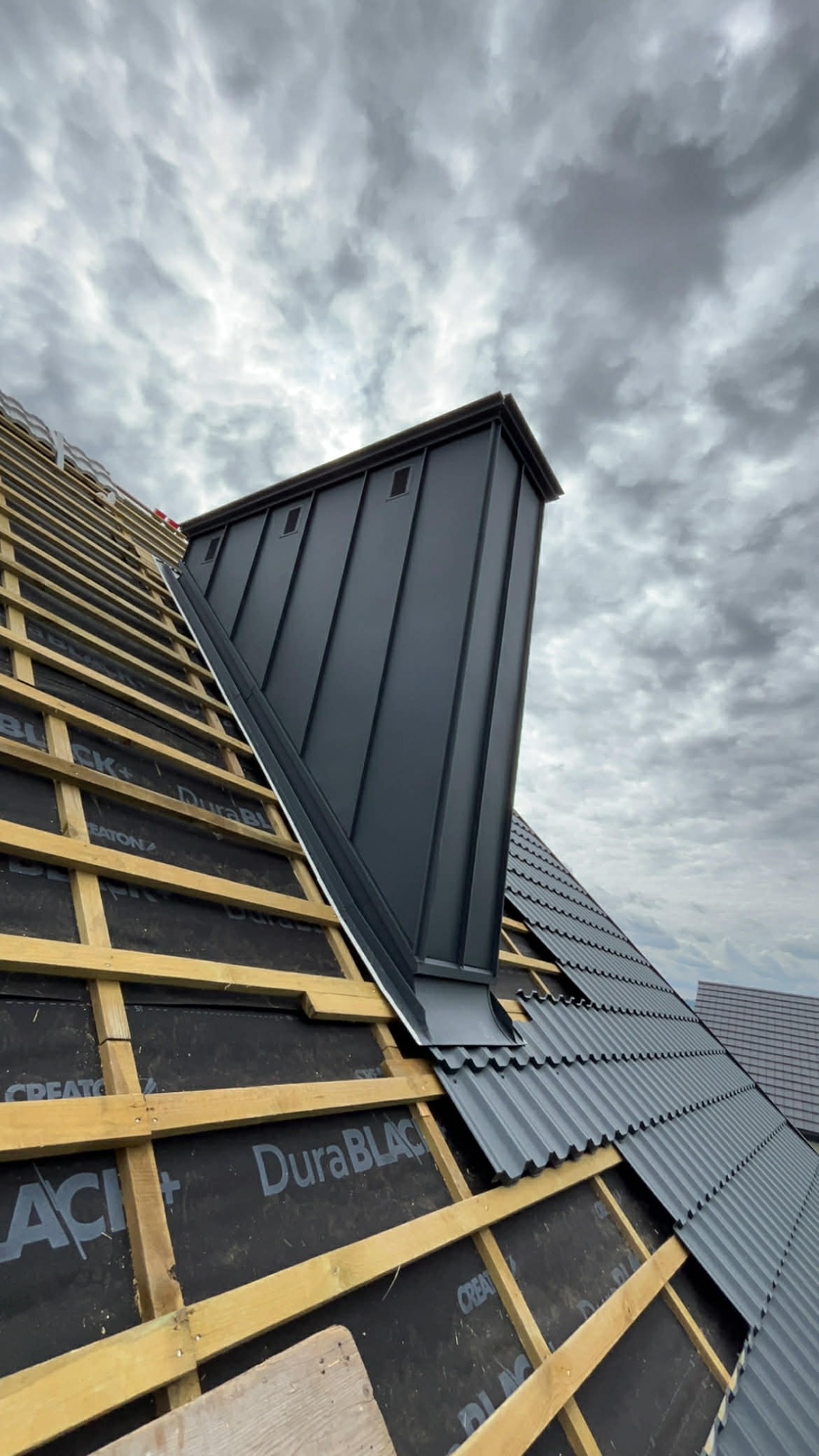 Montaż grafitowej obróbki komina na dachu z drewnianą konstrukcją i membraną, częściowo pokrytego szarą blachodachówką na tle pochmurnego nieba.