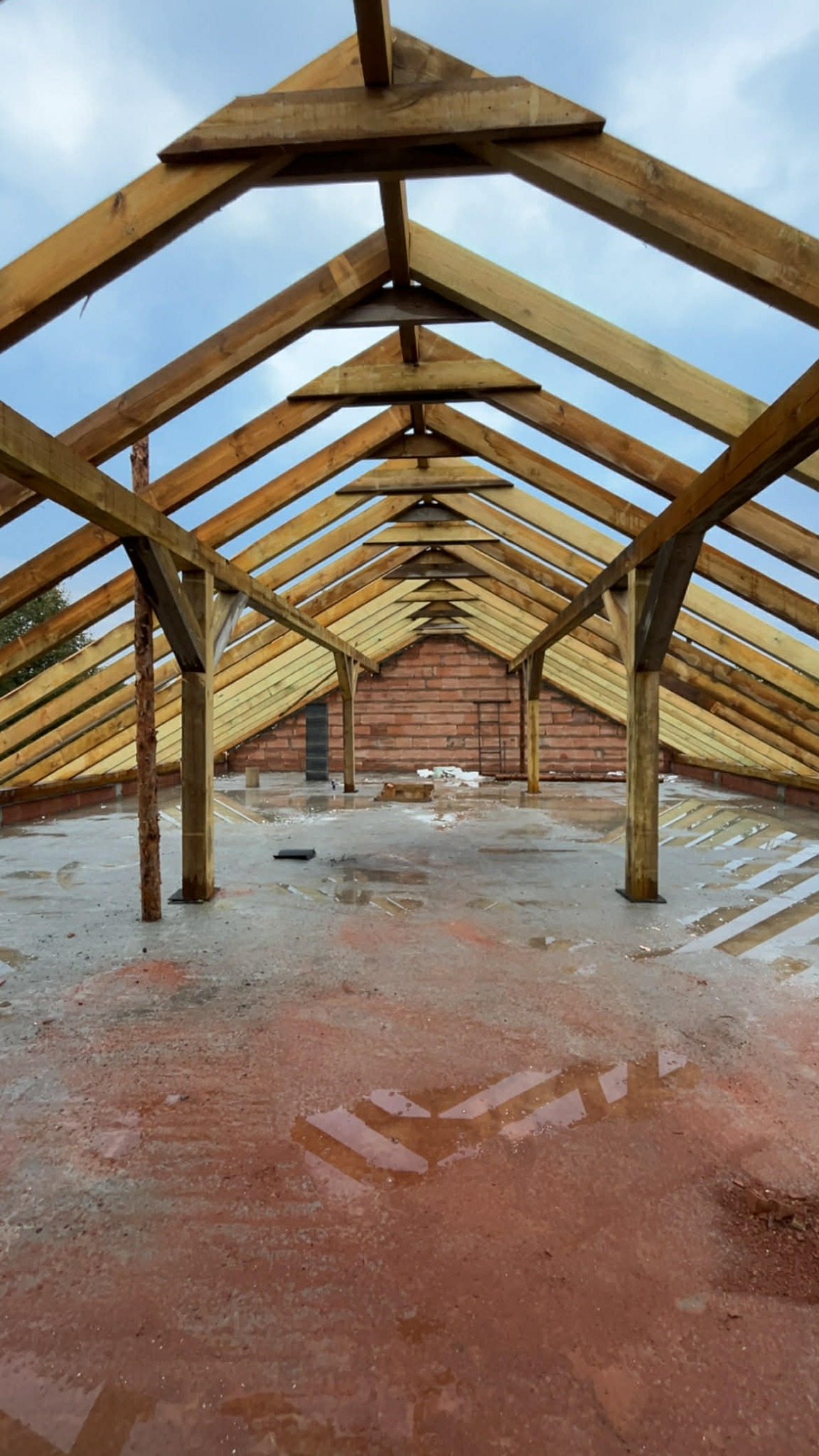 Surowa konstrukcja drewniana dachu z widocznymi belkami i słupami, na tle ceglanej ściany i pochmurnego nieba. Widok z wnętrza budynku w trakcie budowy.