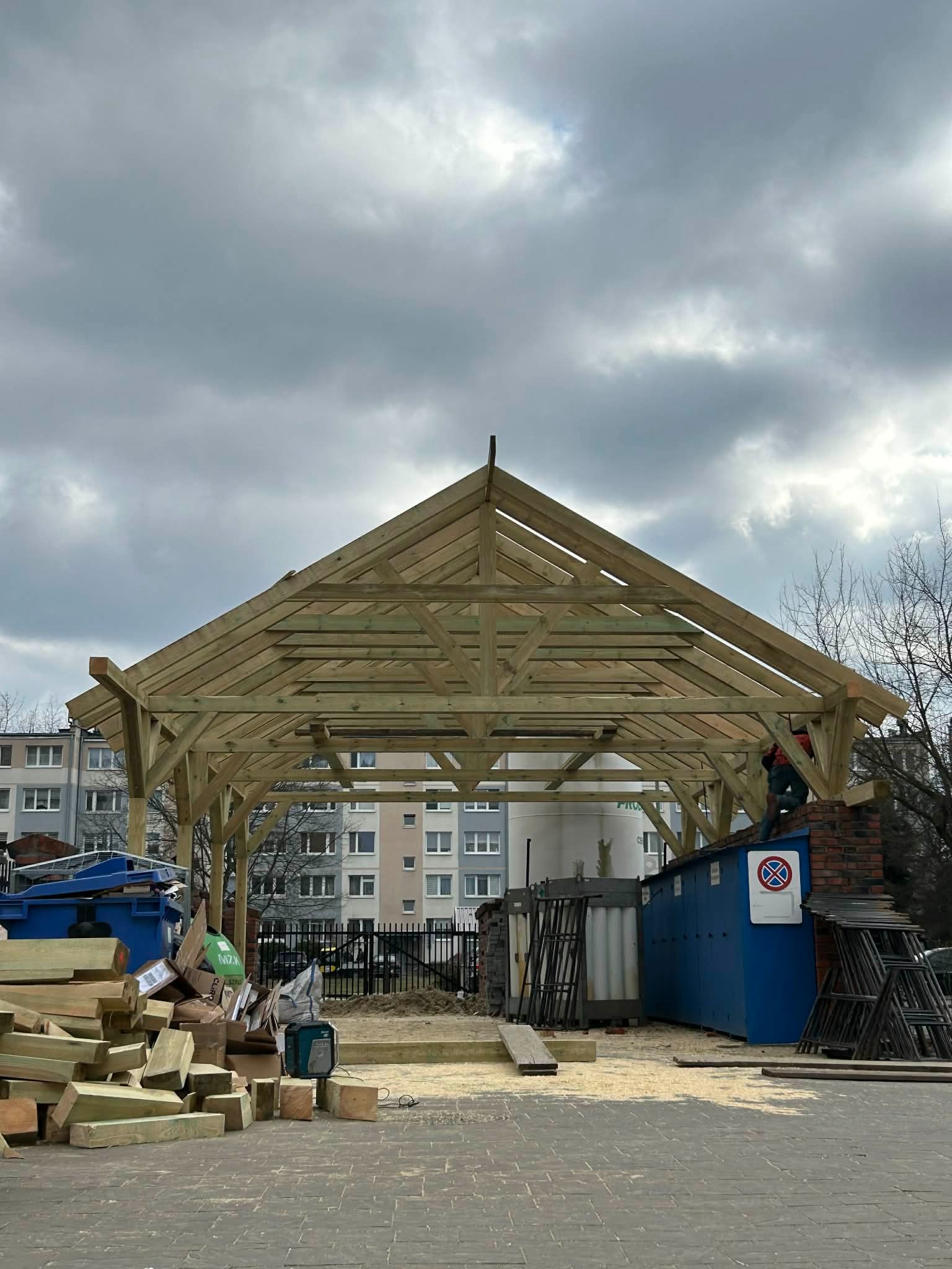 Wznoszenie drewnianej konstrukcji dachu altany na tle pochmurnego nieba i zabudowy miejskiej. Widoczne elementy więźby dachowej i materiały budowlane.