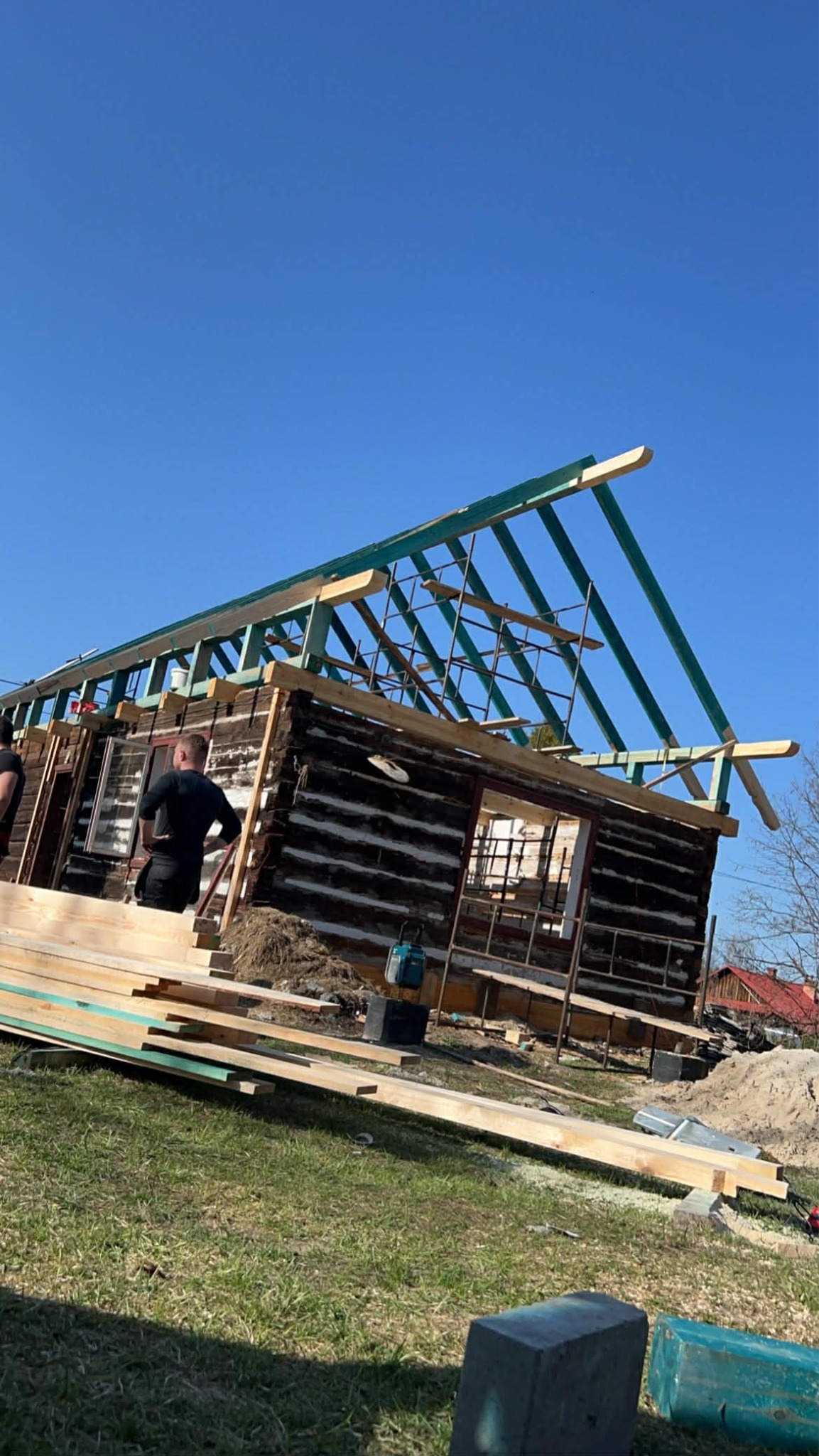 Wznoszenie drewnianej konstrukcji domu z bali, widoczna więźba dachowa w trakcie montażu, deski i narzędzia na trawie, błękitne niebo w tle.