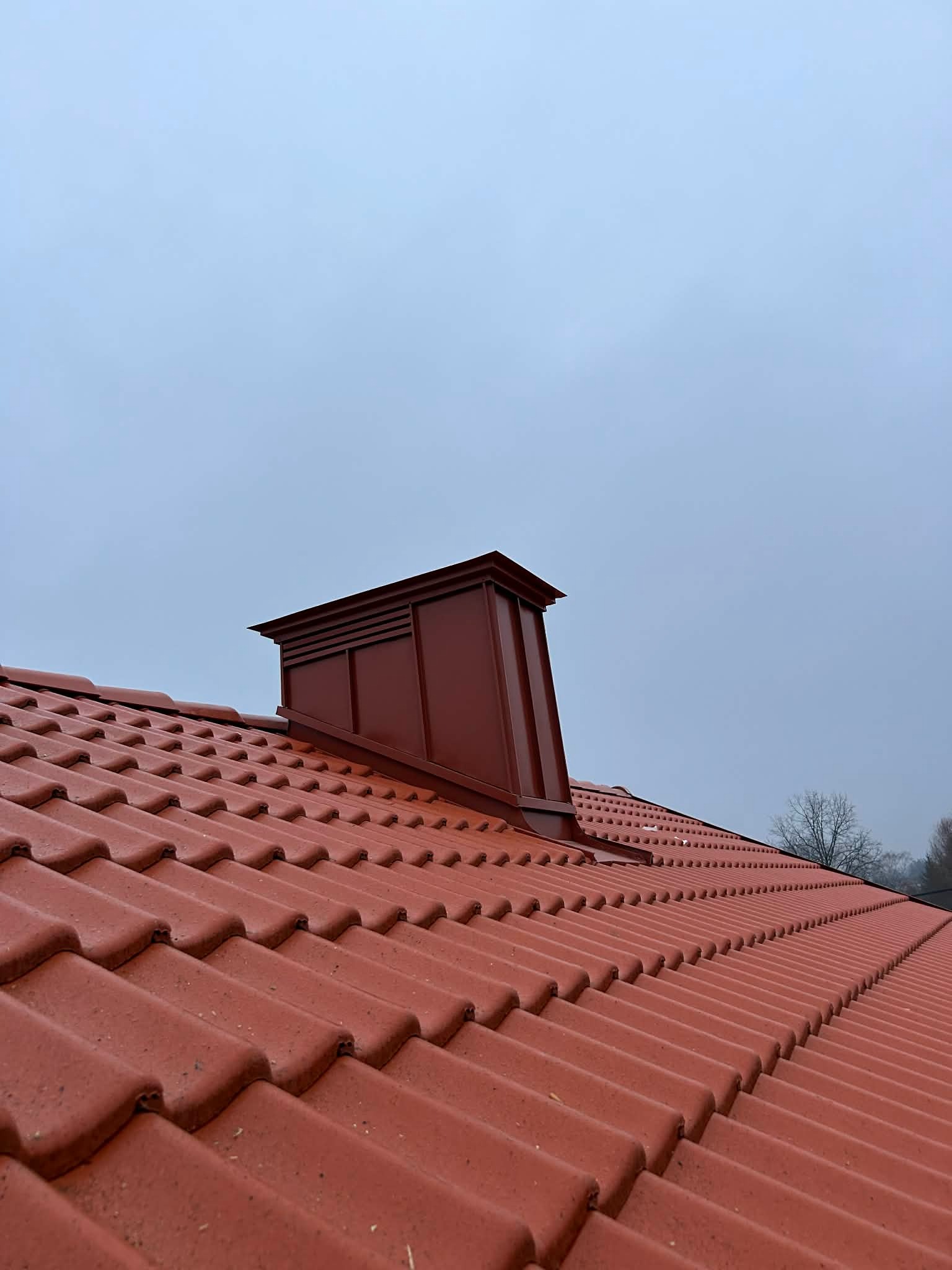 Czerwona dachówka ceramiczna z kominem wentylacyjnym w kolorze, na tle pochmurnego nieba. Widok z dołu, podkreślający regularność ułożenia dachówek.