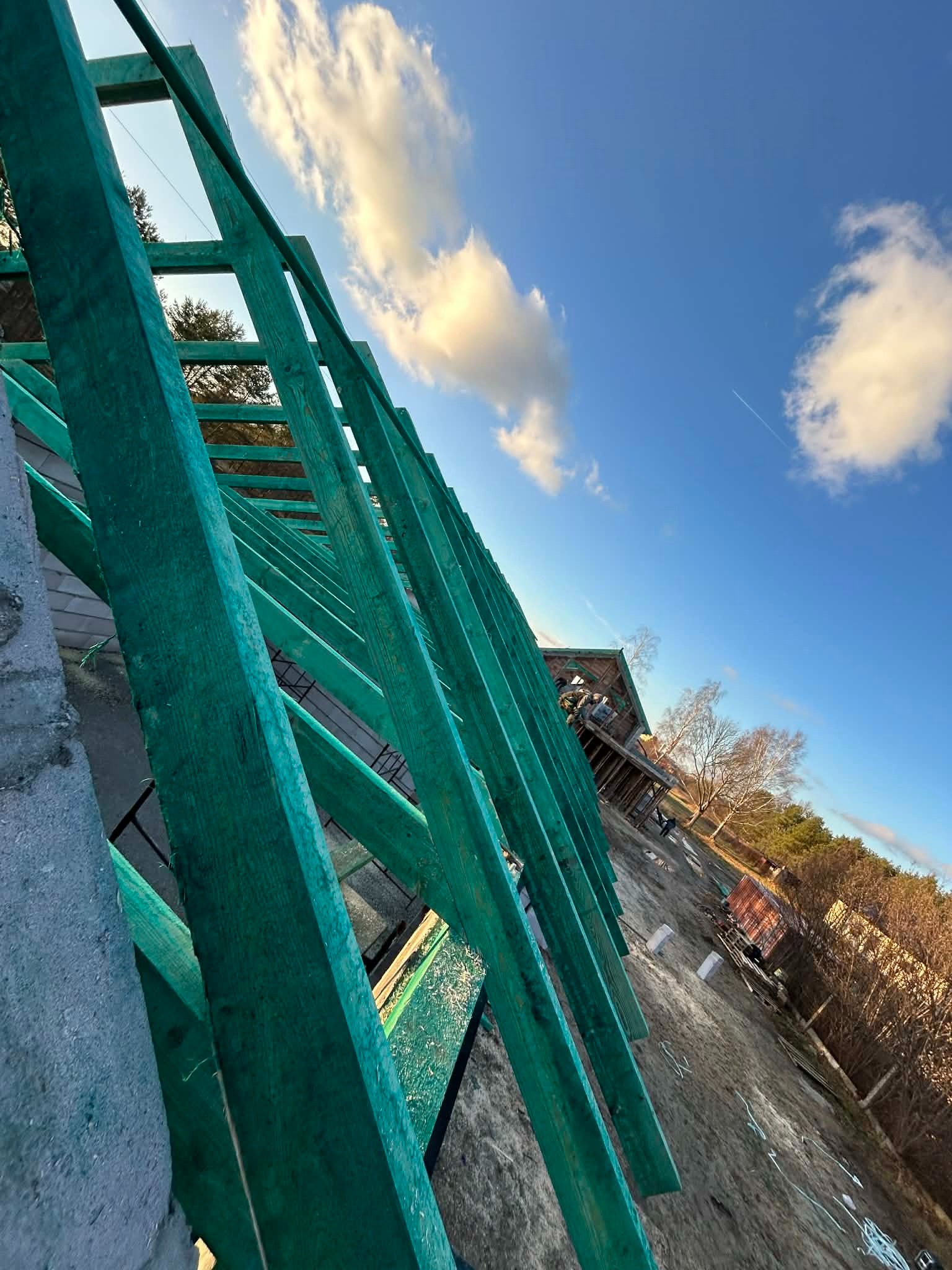 Wznoszenie zielonej więźby dachowej na tle błękitnego nieba z chmurami. Widok z dołu, ukośny kadr. W tle budynek i drzewa.