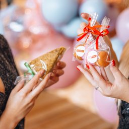 CoZaFoto - Dwie osoby w Krakowie trzymają w dłoniach zapakowane upominki: trójkątną przekąskę z zieloną wstążką oraz woreczki z małymi ciasteczkami, przewiązane czerwoną wstążką i opatrzone okrągłą etykietą...