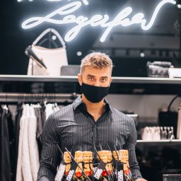 CoZaFoto - Kelner w masce trzyma tacę z mini butelkami szampana Moet z etykietami Karl Lagerfeld na tle sklepu odzieżowego z neonem Karl Lagerfeld.