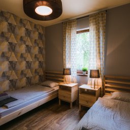 CoZaFoto - Pokój z dwoma łóżkami pojedynczymi, drewnianymi szafkami nocnymi z lampkami i geometryczną tapetą w odcieniach szarości i złota.