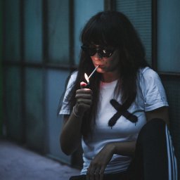 CoZaFoto - Kobieta w okularach przeciwsłonecznych, siedząca na tle szklanej ściany, zapala papierosa zapalniczką; na białej koszulce czarny znak X z efektem kapania.