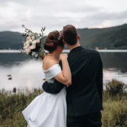 Para młoda, tyłem, obejmująca się na tle jeziora i g&oacute;r; panna młoda trzyma bukiet białych r&oacute;ż i ma ozdobę we włosach.