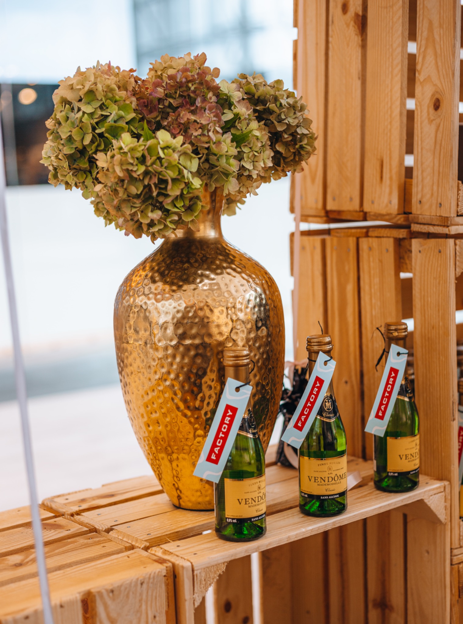 Złoty, teksturowany wazon z hortensjami w odcieniach zieleni i bordo, ustawiony obok trzech butelek bezalkoholowego szampana Vendome Mademoiselle na drewnianej konstrukcji z palet.