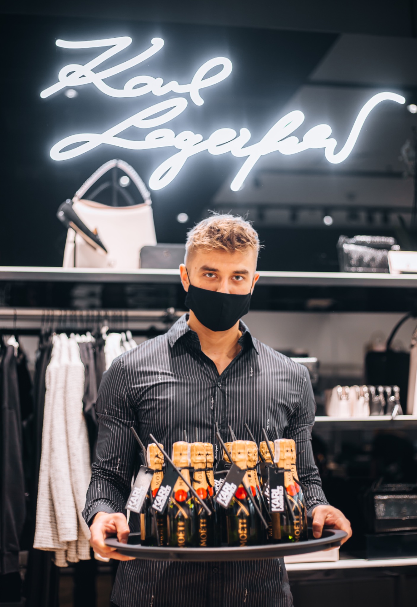 Kelner w masce trzyma tacę z mini butelkami szampana Moet z etykietami Karl Lagerfeld na tle sklepu odzieżowego z neonem Karl Lagerfeld.