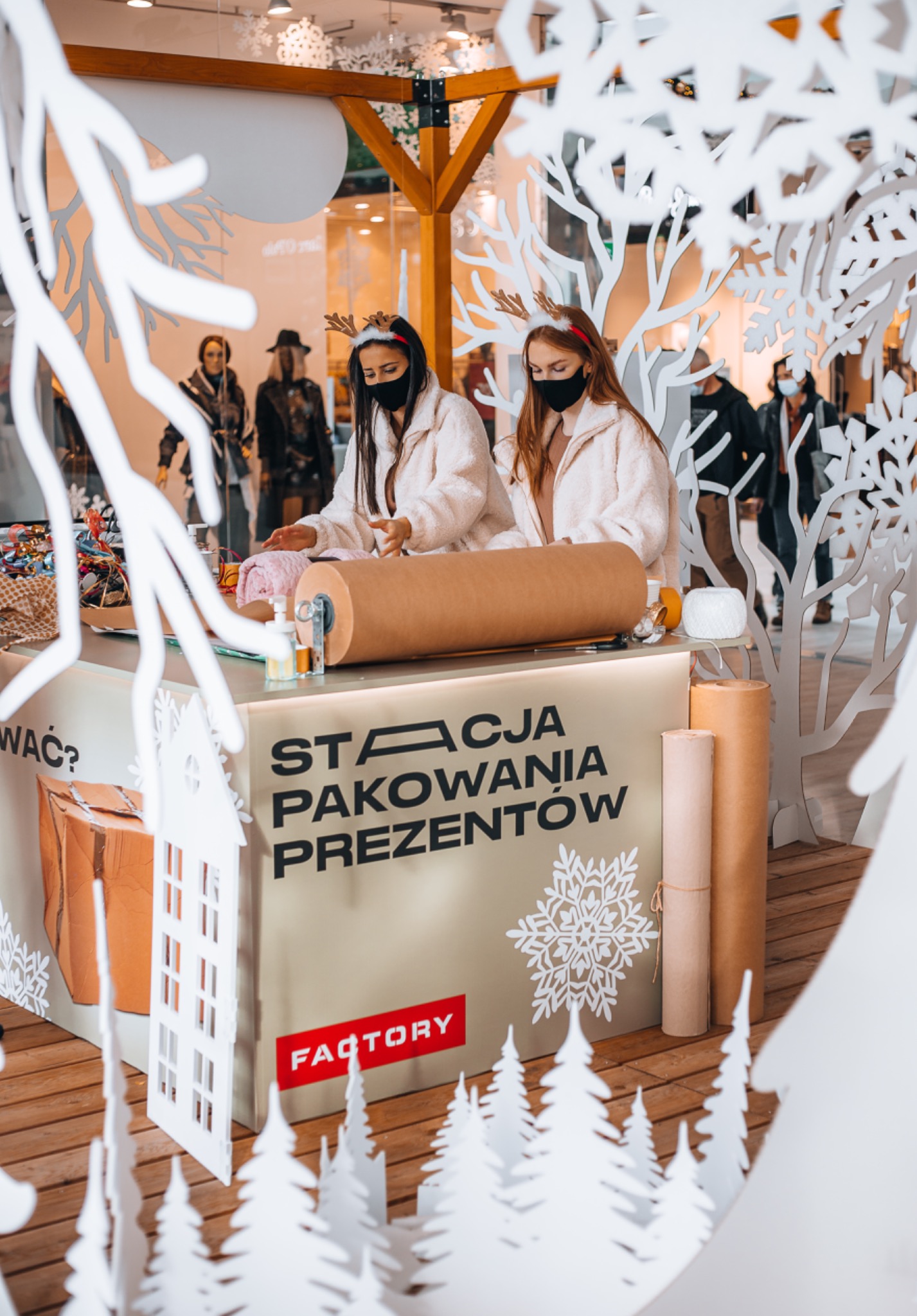 Dwie kobiety w maskach i opaskach z rogami renifera pakują prezenty przy stanowisku z napisem 'Stacja Pakowania Prezentów' w centrum handlowym, widoczne dekoracje świąteczne i przechodnie w tle.